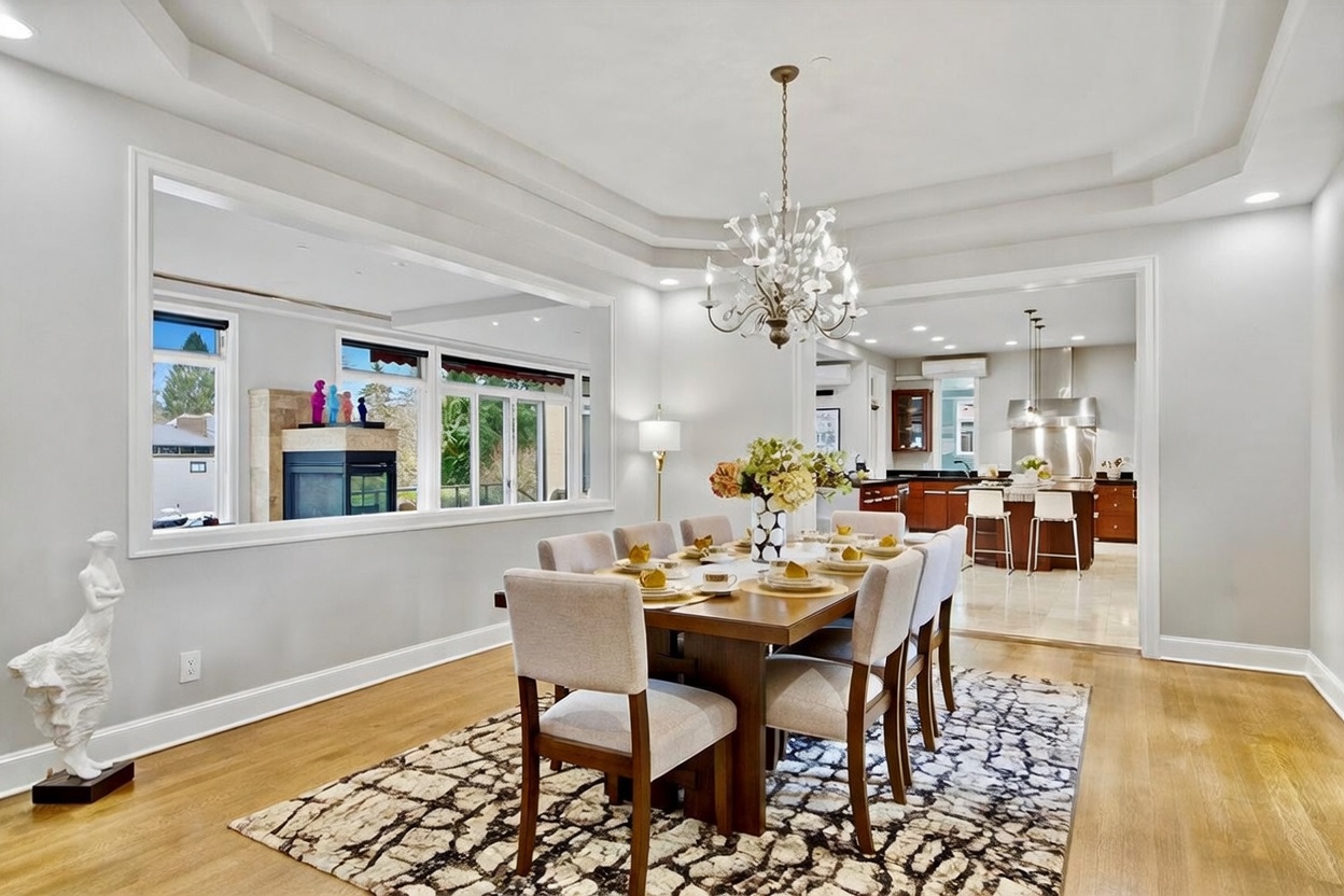 This elegant dining room features a central wooden table surrounded by six upholstered chairs, set atop a large patterned area rug. A crystal chandelier hangs from the tray ceiling, while a large rectangular opening provides a view into the adjacent kitchen and outdoor space. The room is decorated with a tall white sculpture on the left and a gold-framed painting on the wall, creating a sophisticated and inviting atmosphere.