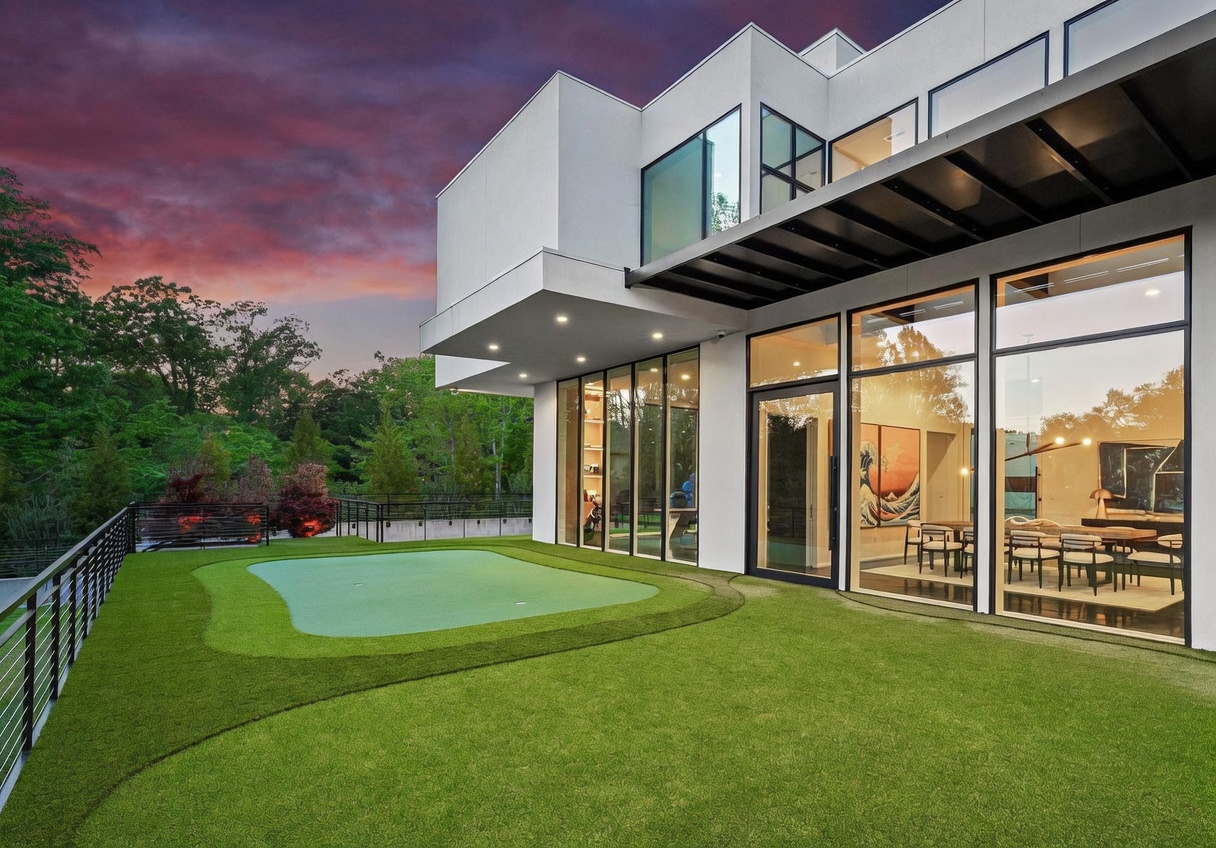 This modern, multi-level home features a spacious elevated terrace equipped with a private putting green. The exterior showcases clean, minimalist lines with large glass panels that provide a seamless transition between the indoor dining area and the outdoor space. The scene is captured at dusk, with the warm interior lighting contrasting against the dramatic, colorful sunset sky.