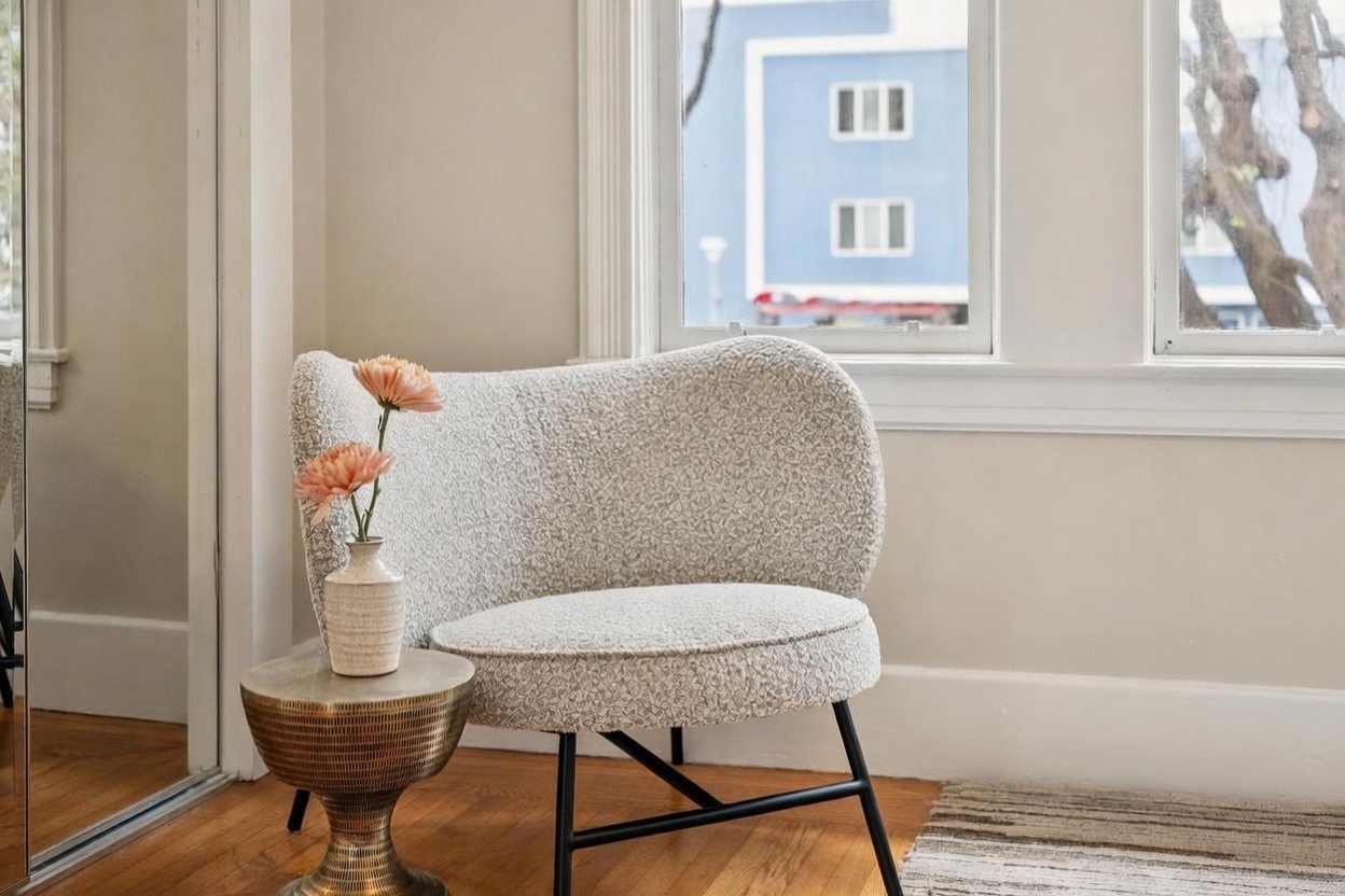 This cozy corner features a textured, light-colored accent chair with a curved back, positioned next to a metallic, drum-style side table holding a small vase with flowers. The space is brightened by large windows that offer a view of the neighborhood, highlighting the warm wood flooring. The overall aesthetic is clean, modern, and inviting, creating a peaceful reading nook or seating area.