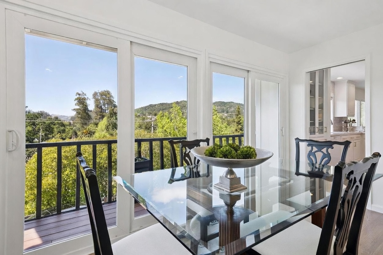 This bright dining area features a glass-topped table with dark-framed chairs, centered in front of a wall of large windows that provide an expansive view of the surrounding trees and hills. The room is finished in crisp white, creating a clean and airy atmosphere that flows into an adjacent kitchen visible through a doorway. The perspective captures the seamless transition between the indoor dining space and the outdoor balcony, emphasizing the home's connection to the natural landscape.