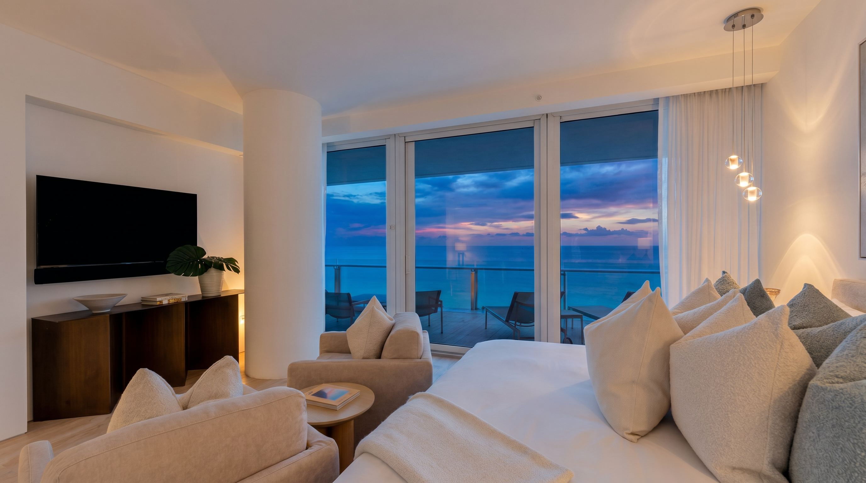This bright, contemporary bedroom features a plush bed with neutral-toned pillows and a seating area with two armchairs facing a wall-mounted television. The room is defined by expansive floor-to-ceiling windows that provide an unobstructed, panoramic view of the ocean and a private balcony. The minimalist aesthetic is complemented by light wood-toned flooring and elegant pendant lighting, creating a serene and luxurious atmosphere.