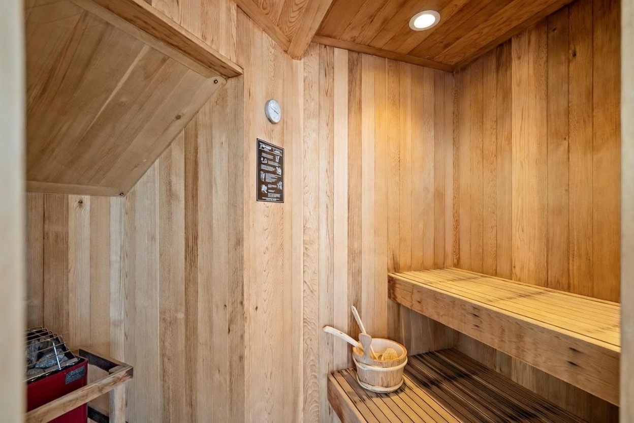 This image showcases a compact, wood-paneled sauna room designed for relaxation. The space features tiered wooden benches, a traditional sauna heater with stones, and a wooden bucket with a ladle, all enclosed in warm-toned wood planks. A small thermometer is mounted on the wall, and the room is illuminated by a recessed ceiling light, creating a cozy and inviting atmosphere.