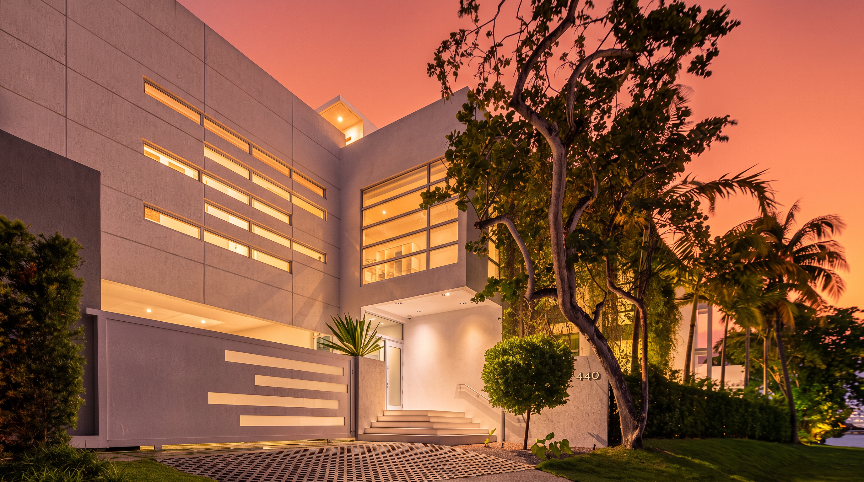 This striking modern residence features a minimalist white facade with geometric horizontal window accents and a prominent multi-story glass entryway. The property is framed by a mature tree and lush landscaping, with a paved driveway leading to a set of steps that ascend to the front door. Captured at twilight, the home is illuminated by warm exterior lighting, creating a sophisticated and inviting atmosphere.