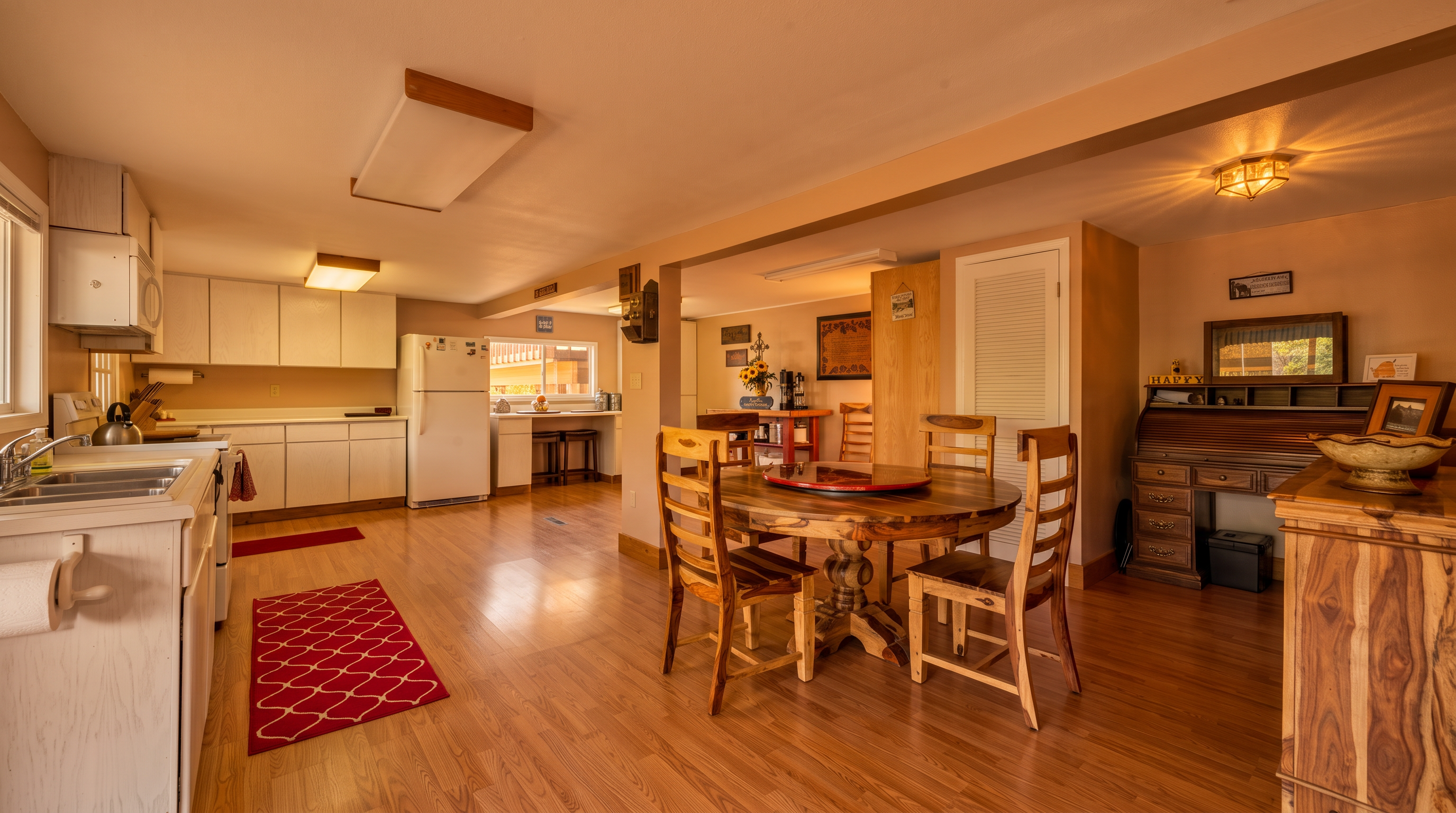 This spacious, open-concept kitchen and dining area features warm wood-look flooring and white cabinetry paired with a large, rustic circular wooden dining table. The kitchen is well-equipped with full-sized appliances, including a white refrigerator and range, while the adjacent office corner includes a traditional roll-top desk, creating a functional multi-purpose space. With its bright lighting and connected layout, the home offers a welcoming and comfortable atmosphere perfect for family living and entertaining.