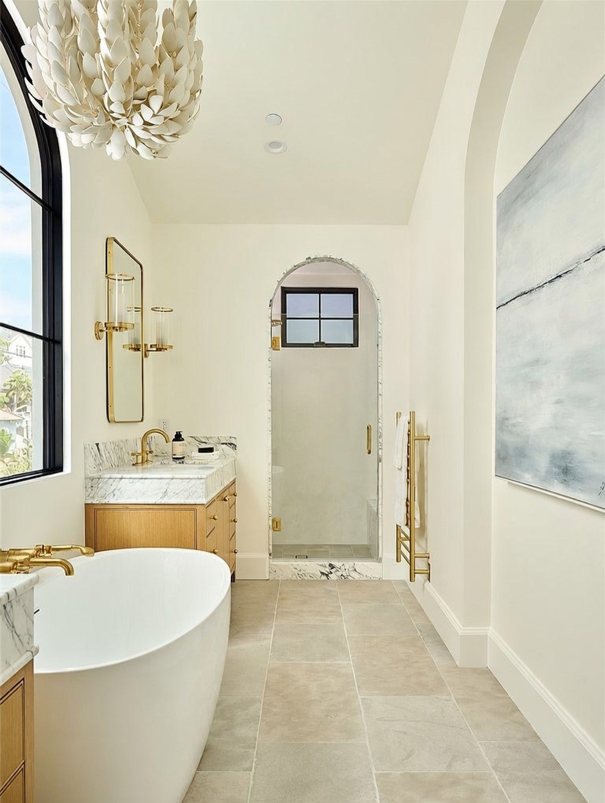 This elegant primary bathroom features a freestanding soaking tub in the foreground and a walk-in shower with an arched entry in the background. The space is accented by a light wood vanity with a stone countertop, a gold-framed mirror, and a decorative chandelier. The neutral color palette and clean lines create a sophisticated and serene atmosphere.