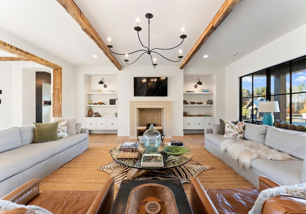 This spacious living room features a symmetrical layout with two large light-gray sofas facing each other across a glass-topped coffee table. A central fireplace with a stone surround serves as the focal point, flanked by built-in shelving units and wall-mounted sconces. The room is accented by exposed wooden ceiling beams, a textured rug over light wood flooring, and large windows that provide ample natural light and a view of the outdoors.