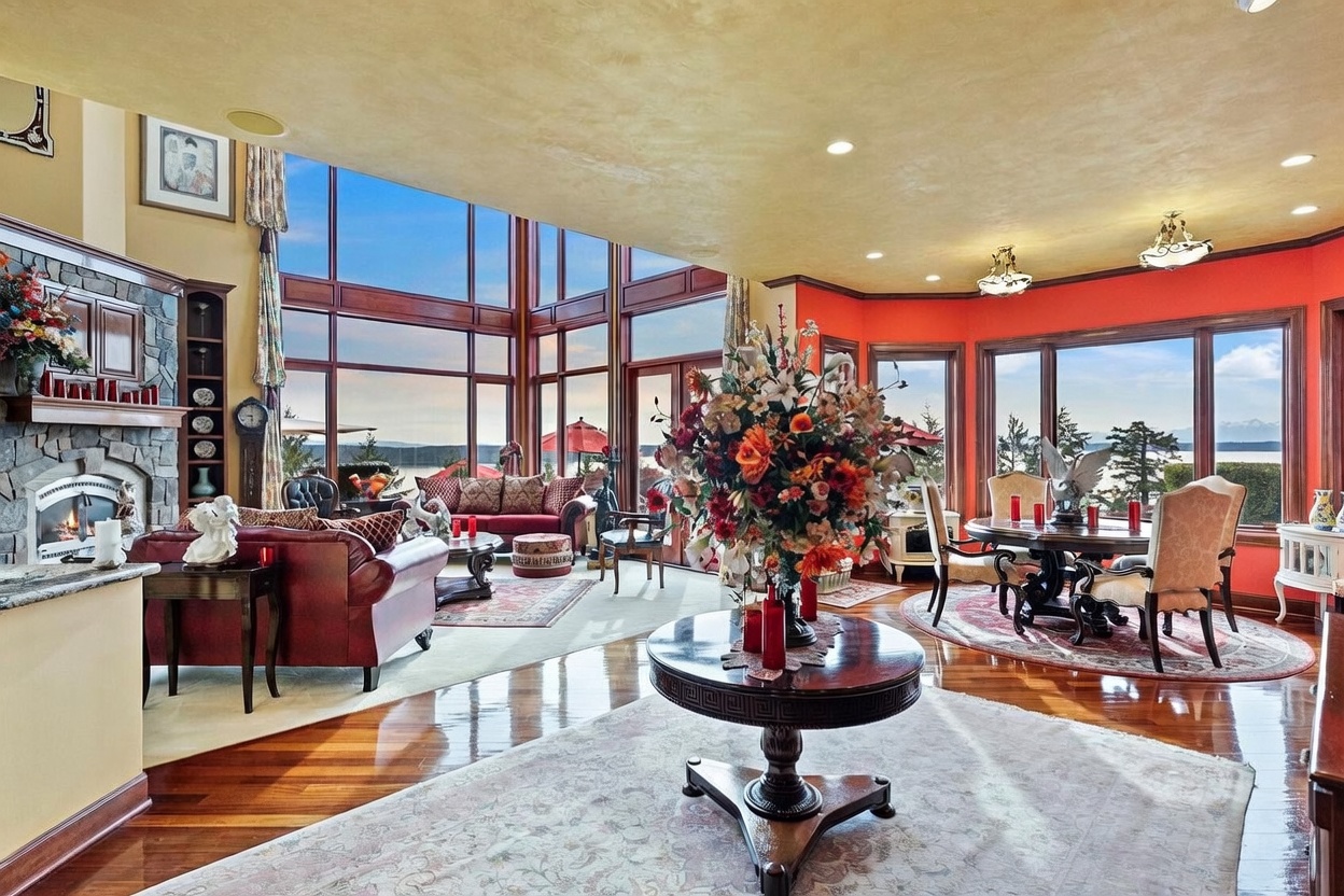 This expansive, open-concept living and dining area features rich hardwood flooring and a warm, golden-toned ceiling. The space is anchored by a stone-clad chimney breast with a fireplace and a large, central circular table topped with a vibrant floral arrangement. Massive windows provide panoramic views of the water and distant mountains, creating a bright and airy atmosphere that blends luxury with natural scenery.