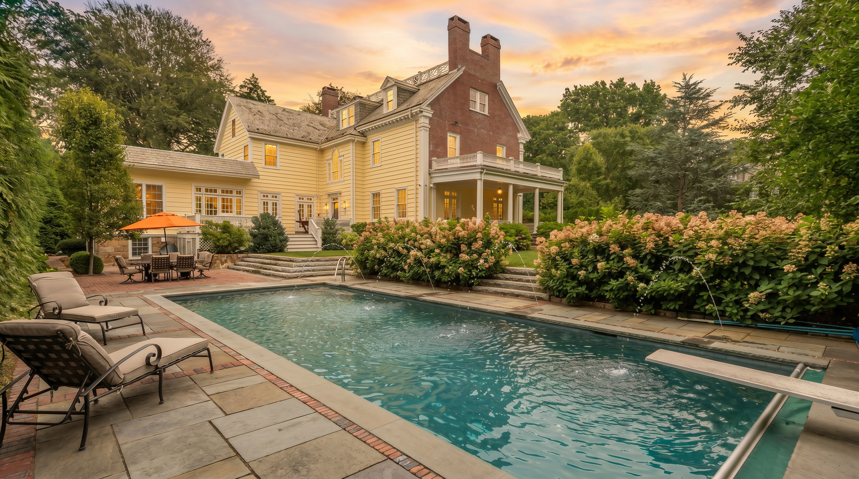 This rear exterior view showcases a classic, multi-story home with yellow siding and a prominent brick chimney, set against a lush, mature landscape. A rectangular swimming pool with a diving board serves as the focal point, complemented by a stone-paved patio area featuring lounge chairs and an outdoor dining set under a bright orange umbrella. The scene is framed by vibrant hydrangea bushes and manicured greenery, creating a serene and inviting backyard retreat.