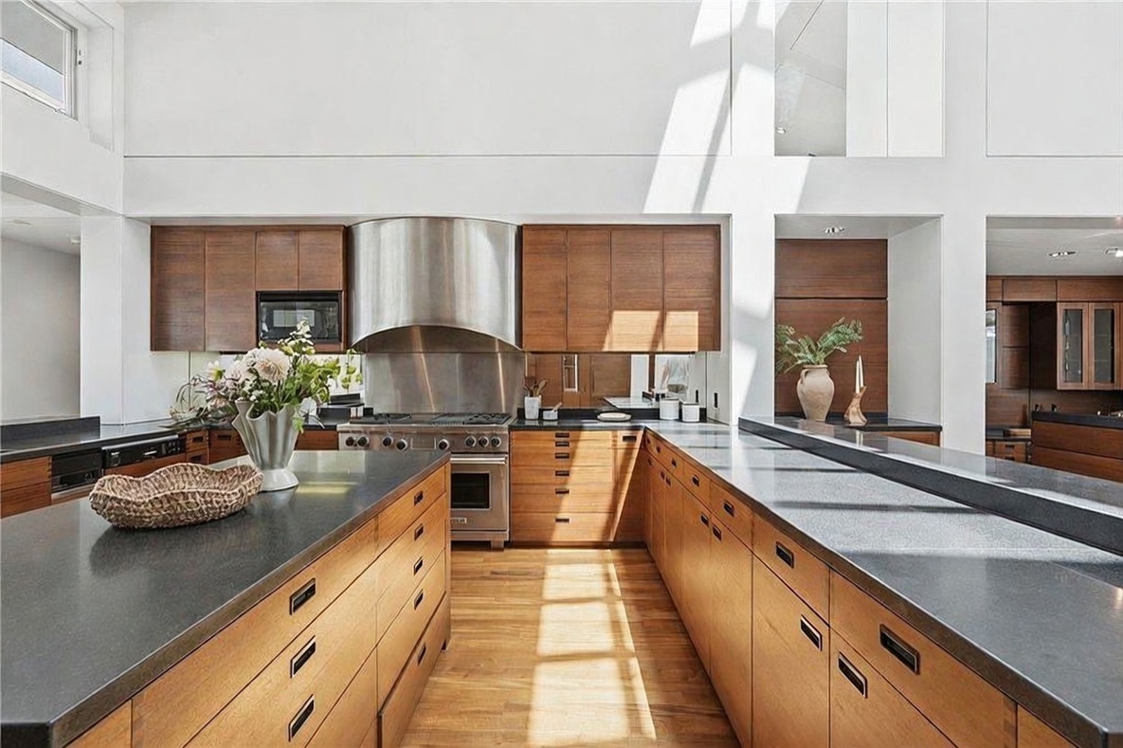 This modern kitchen features warm-toned wood cabinetry paired with dark stone countertops and a large central island. A stainless steel range with a prominent vent hood serves as the focal point, while the high ceilings and open layout create an airy, expansive atmosphere. The space is bathed in natural light, highlighting the clean lines and contemporary design aesthetic.
