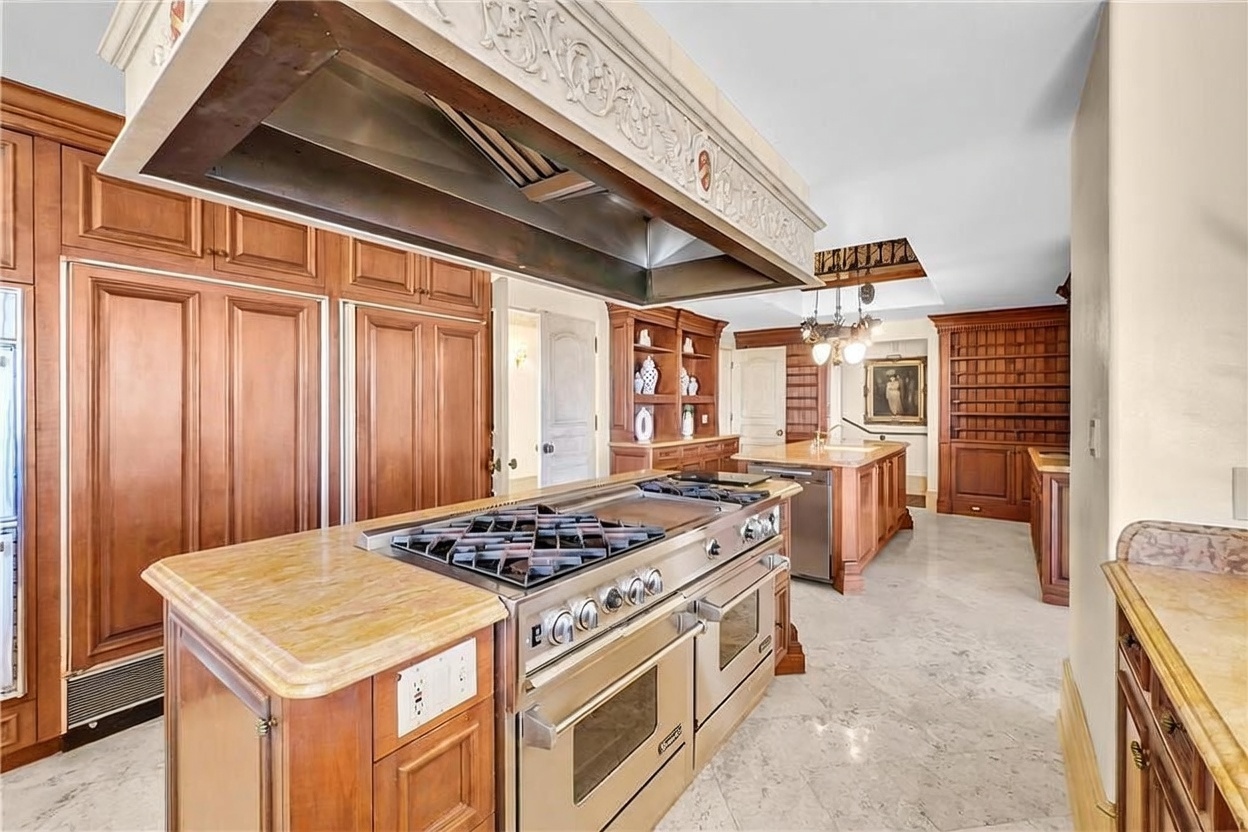 This expansive, traditional-style kitchen features a large central island equipped with a professional-grade stainless steel range and a decorative, oversized vent hood above. The space is defined by rich, warm-toned cabinetry, light-colored stone countertops, and polished stone flooring. A secondary island with a sink and dishwasher sits in the background, complemented by built-in shelving and a classic chandelier, creating a sophisticated and functional culinary environment.