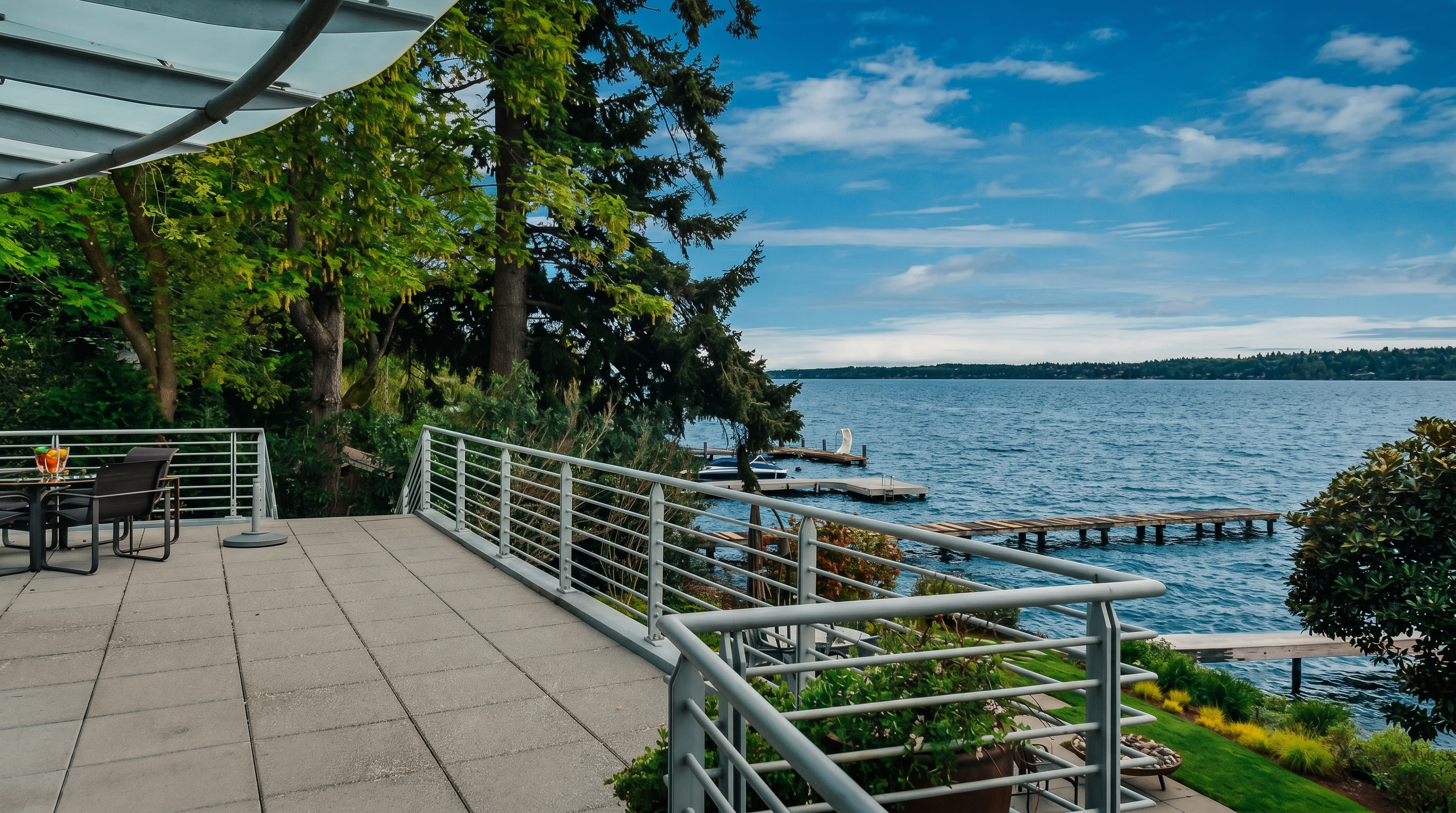 This expansive, elevated deck offers a panoramic view of a large lake, featuring a modern metal railing and paved stone flooring. The space includes a small outdoor dining set and provides a seamless transition to the lush, green lawn below, which leads directly to a private dock. The scene is captured from a slightly elevated perspective, emphasizing the serene waterfront setting and the open, airy atmosphere of the outdoor living area.