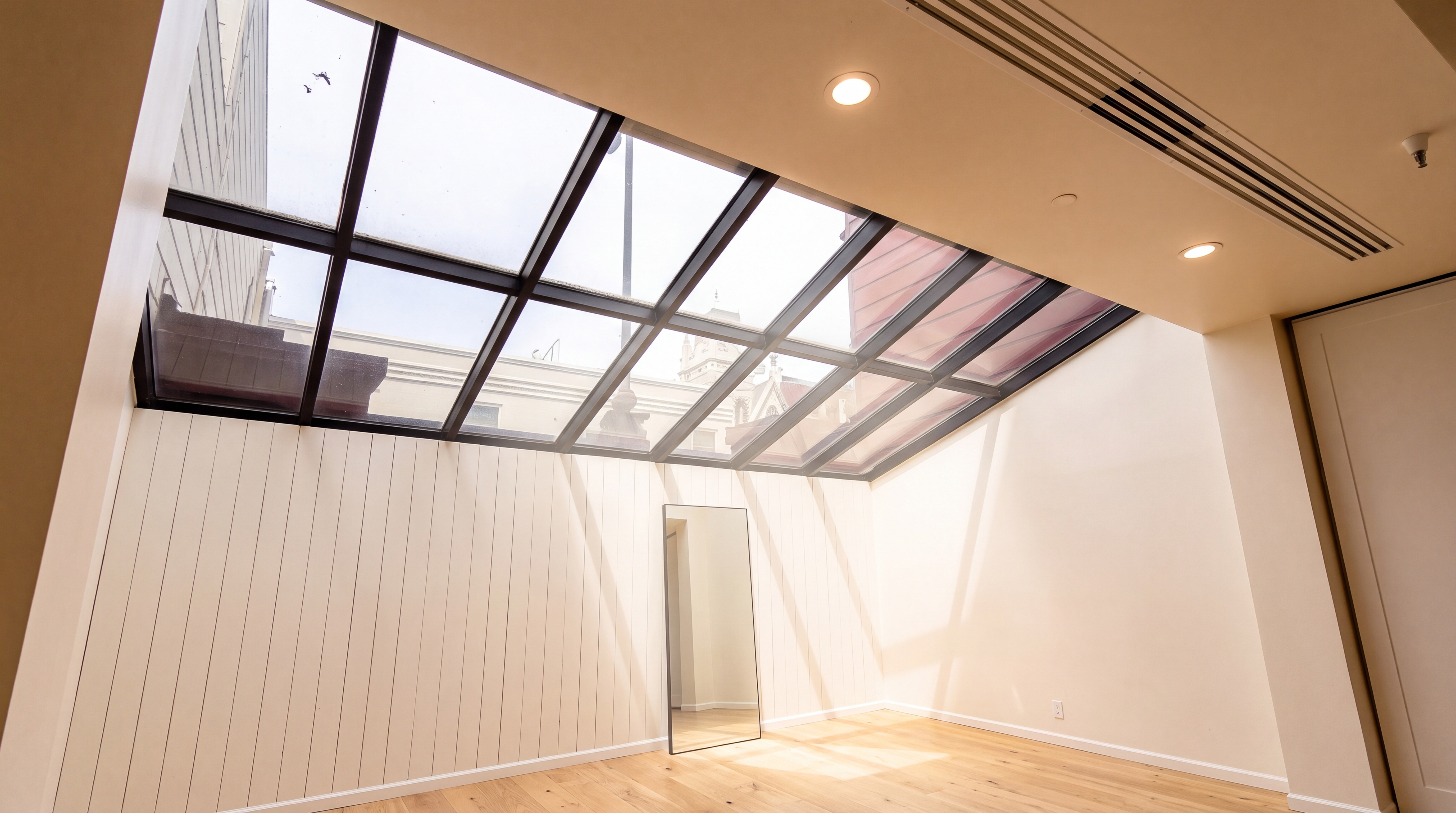 This sun-drenched room features a prominent angled glass skylight ceiling that creates an airy and open architectural feel. The space is finished with light-toned wooden flooring and panelled walls, housing a minimalist free-standing mirror as the focal point. It serves as a versatile, light-filled area perfect for a modern living space or an indoor garden.