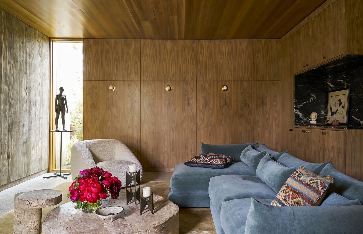 This sophisticated living room features warm wood-paneled walls and a textured, light-colored armchair paired with a plush, blue sectional sofa. A large, circular stone coffee table sits in the center, adorned with a vibrant bouquet of red flowers and decorative candle holders. The space is accented by a tall window showcasing a sculptural figure and a dark, polished stone feature wall with a framed print, creating a refined and artistic atmosphere.