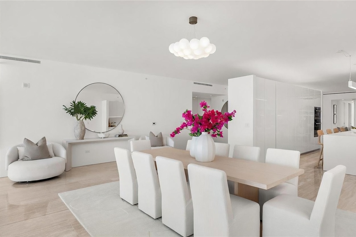 This bright and airy dining room features a long, light-toned wood table surrounded by eight white upholstered chairs, set atop a neutral area rug. A large, modern cloud-like pendant light fixture hangs above the table, while a circular mirror and a console table with a large vase of greenery anchor the adjacent wall. The space is characterized by a minimalist, monochromatic aesthetic with clean white walls and polished light-colored flooring, creating a sophisticated and open atmosphere.