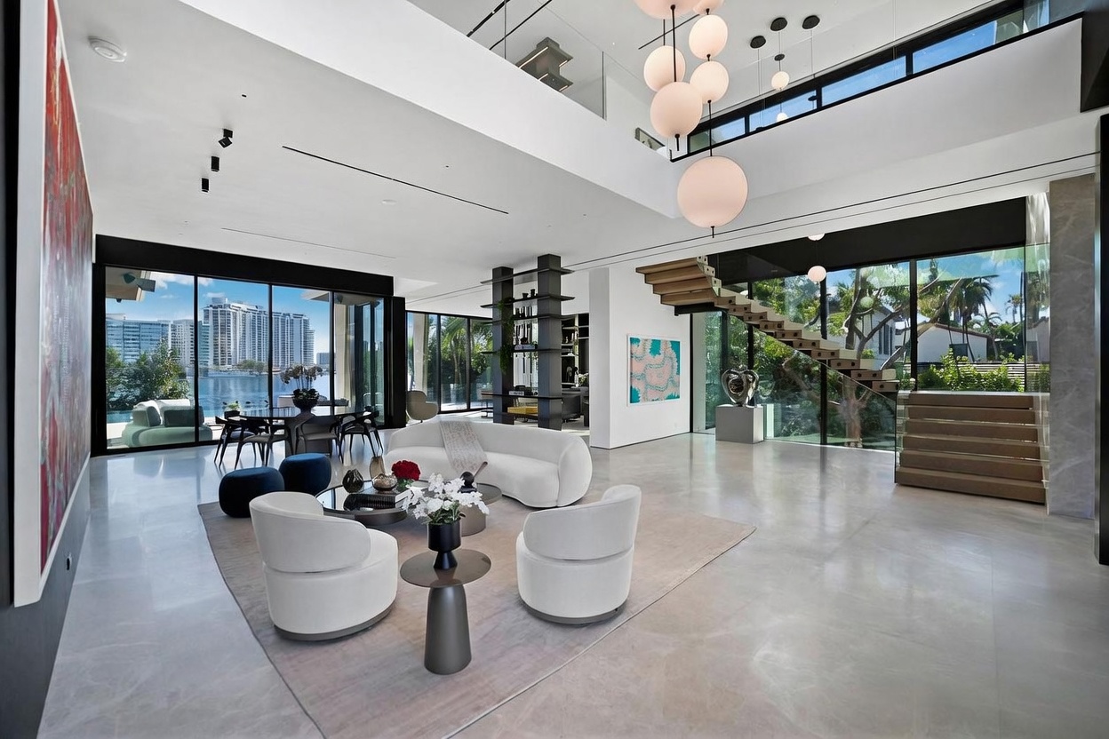 This expansive, double-height living room features a contemporary open-concept design with polished light-toned flooring and a striking sculptural staircase. Large glass walls provide panoramic views of the waterfront and lush greenery, while a cluster of spherical pendant lights serves as a dramatic focal point above the seating area. The space is furnished with a curved white sofa and matching armchairs, creating a sophisticated and airy atmosphere.