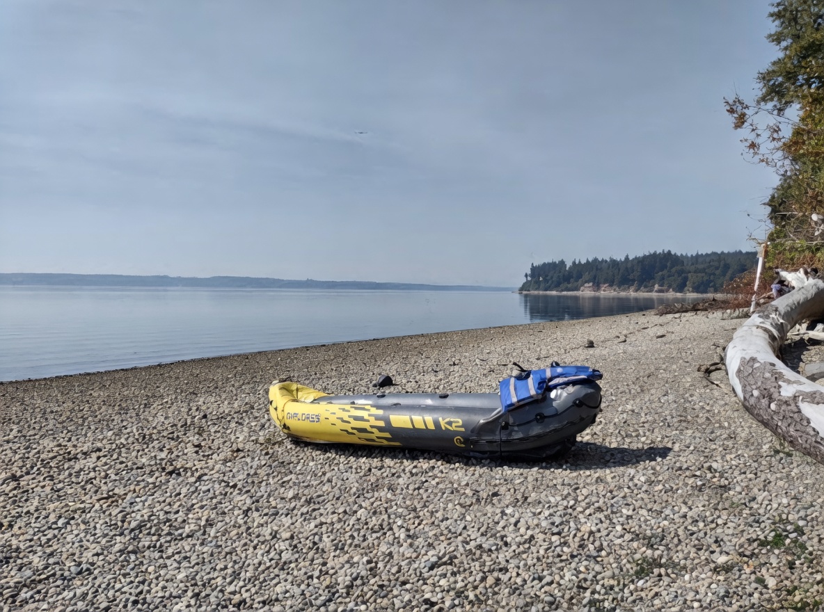 This image captures a serene, pebbled shoreline along a calm body of water, likely a beach or waterfront access point for a community. An inflatable kayak rests on the stones in the foreground, while a large piece of driftwood lies to the right, framing the expansive view of the water and distant treeline. The scene conveys a peaceful, recreational atmosphere typical of a waterfront property or community amenity.