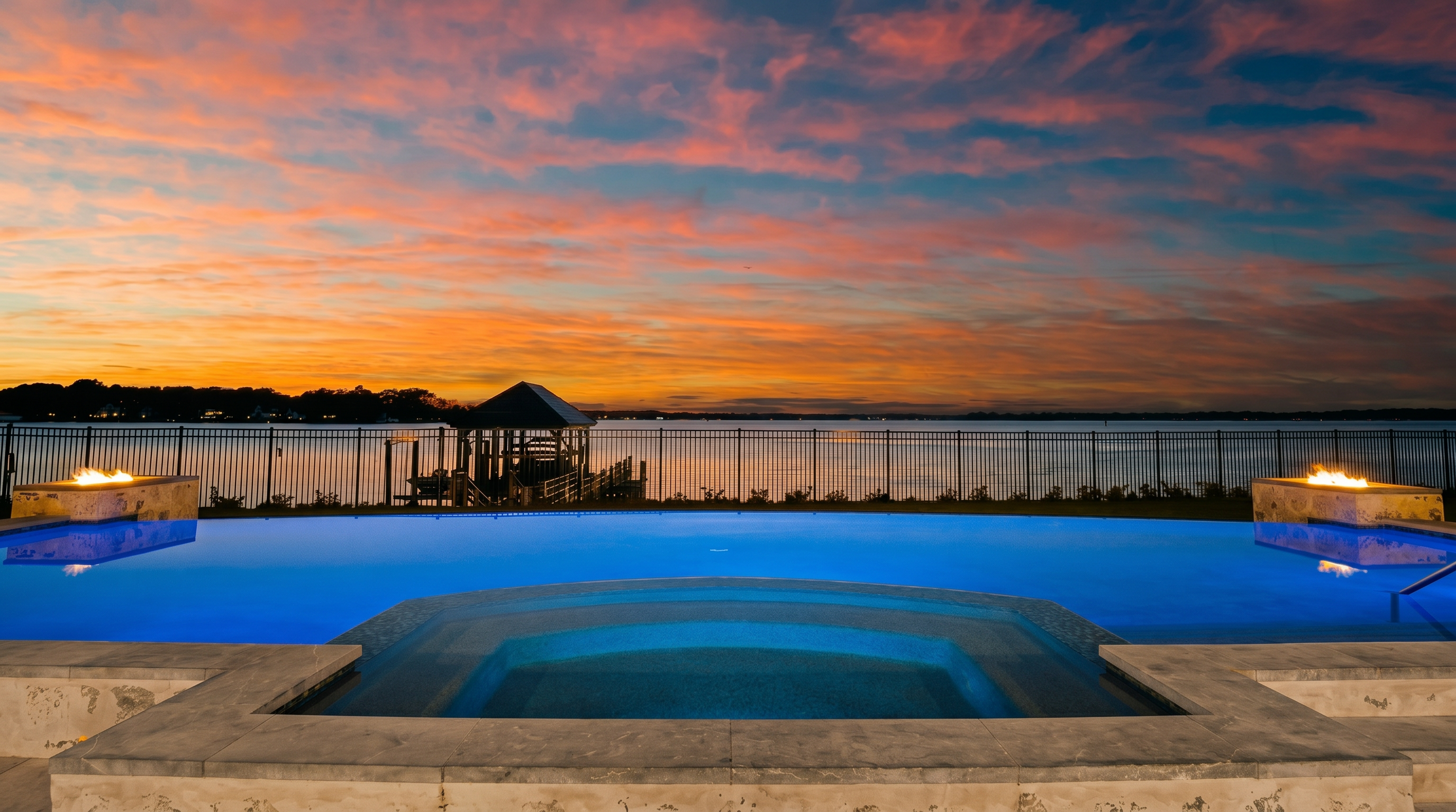 This stunning twilight view captures a luxurious infinity-edge pool and integrated spa overlooking a serene lake. Two stone-clad fire features flank the pool, casting a warm glow that contrasts with the vibrant orange and purple sunset sky. A metal fence separates the pool deck from the water, where a private gazebo sits at the end of a dock, creating a peaceful and high-end atmosphere.