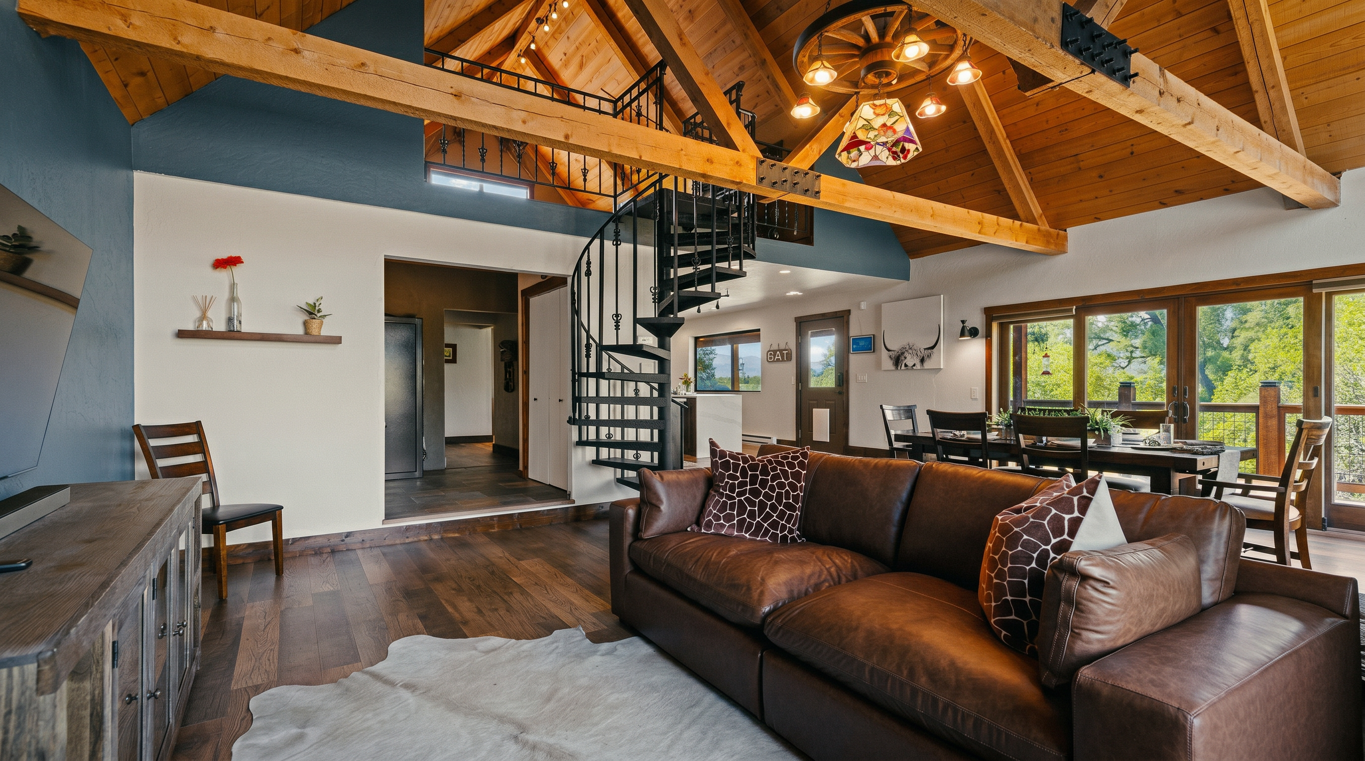 This open-concept living area features a vaulted ceiling with exposed wooden beams and a rustic wagon-wheel chandelier. A large brown leather sofa with patterned throw pillows sits atop a cowhide rug, while a spiral staircase serves as a striking architectural focal point. The space flows seamlessly into a dining area and hallway, with large windows providing ample natural light and views of the surrounding greenery.