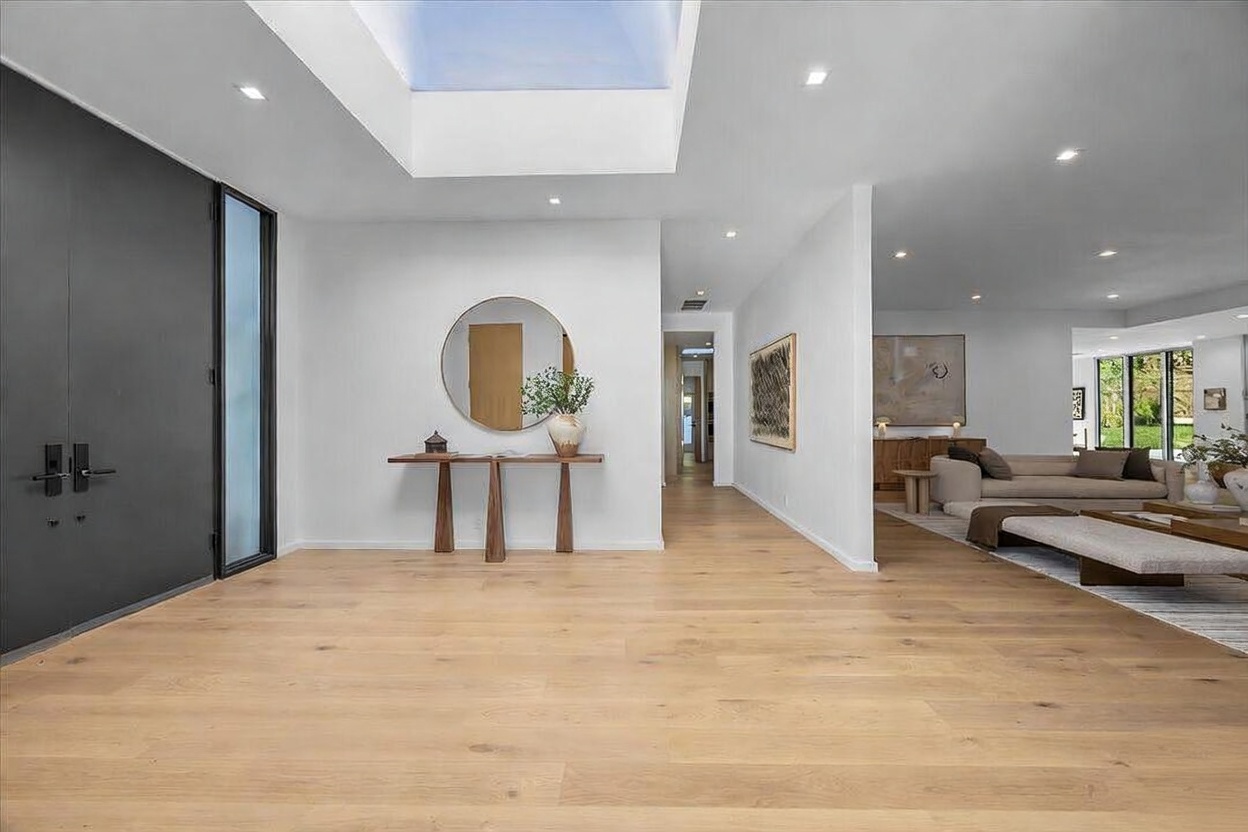 This bright and airy entryway features light wood flooring and a minimalist aesthetic, highlighted by a large skylight that floods the space with natural illumination. A sleek console table sits beneath a circular mirror, while a dark, modern door provides a striking contrast to the clean white walls. The perspective looks from the front door toward a spacious, open-concept living area, emphasizing the home's seamless flow and contemporary design.