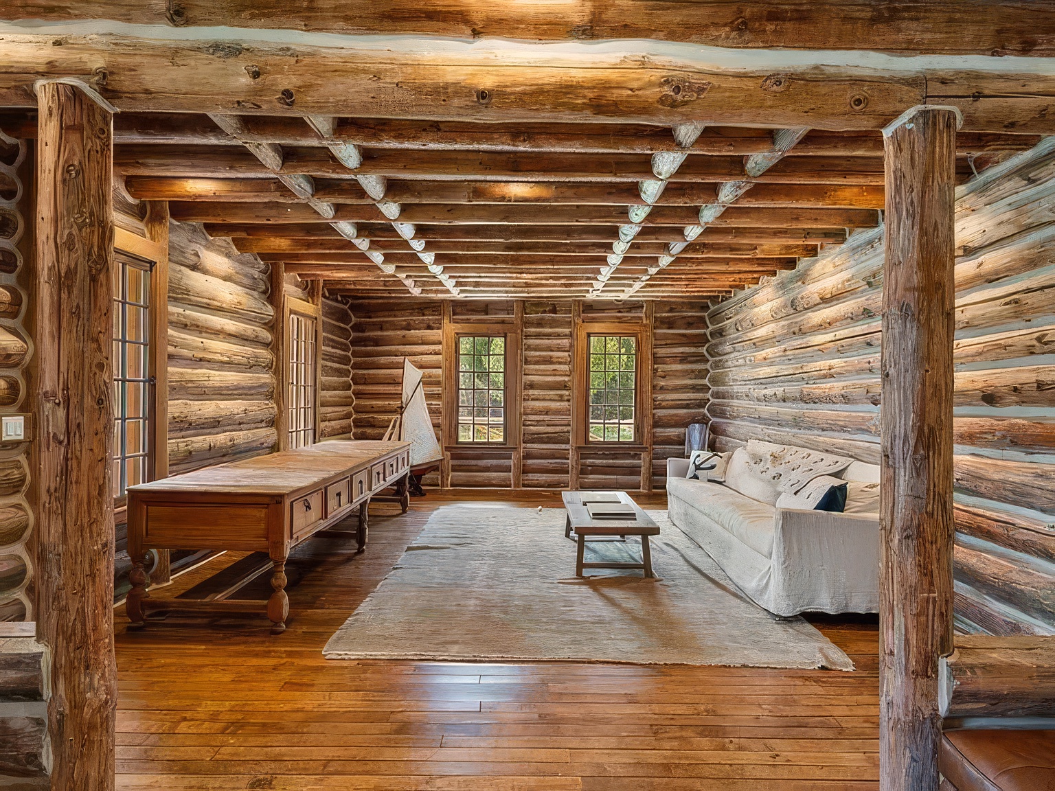 This rustic living room features exposed log walls and a beamed ceiling, creating a warm and authentic cabin aesthetic. The space is furnished with a long wooden table on the left and a comfortable light-colored sofa with a coffee table on the right, all anchored by a large area rug. The perspective is centered, looking through two prominent log pillars into the main seating area, which is illuminated by natural light from the windows on the back wall.