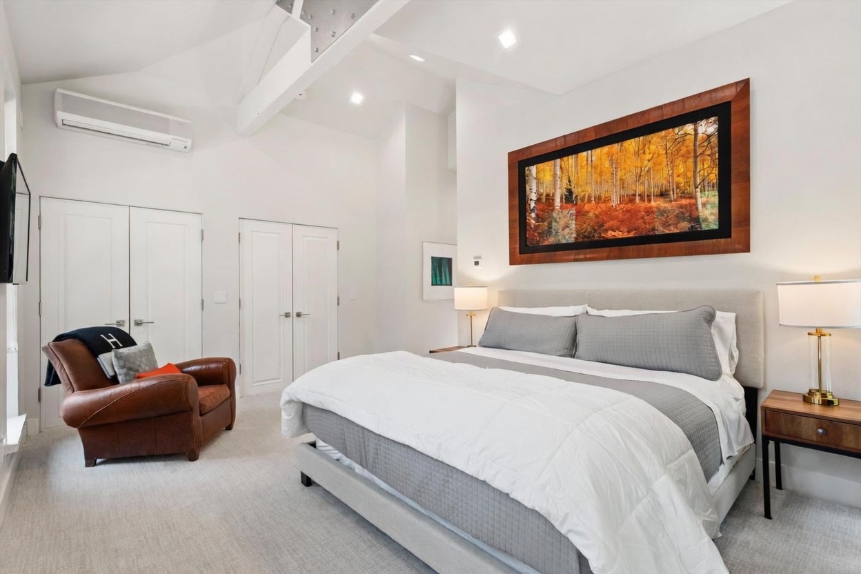 This bright and airy primary bedroom features a vaulted ceiling with exposed structural beams and a neutral color palette. The room is anchored by a large bed with a light-toned upholstered headboard, complemented by a leather armchair in the corner and a large landscape art piece above the bed. The space is well-lit by bedside lamps and recessed lighting, creating a clean, modern, and comfortable atmosphere.