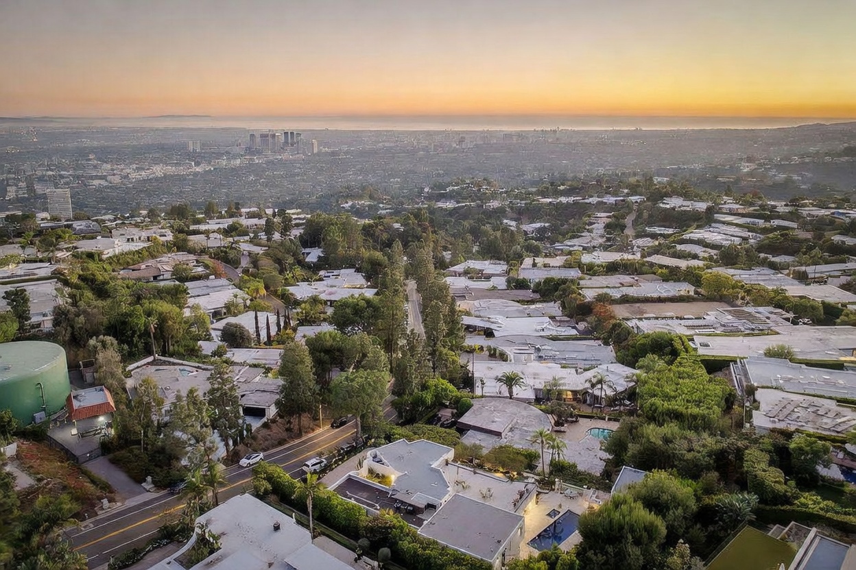 This high-angle aerial view captures a sprawling residential neighborhood nestled in a lush, hilly landscape during a golden sunset. The scene highlights the dense canopy of mature trees interspersed with modern homes, winding roads, and a distant, panoramic view of the city skyline and coastline. The perspective emphasizes the expansive scale of the property and its prime location within a quiet, elevated community.