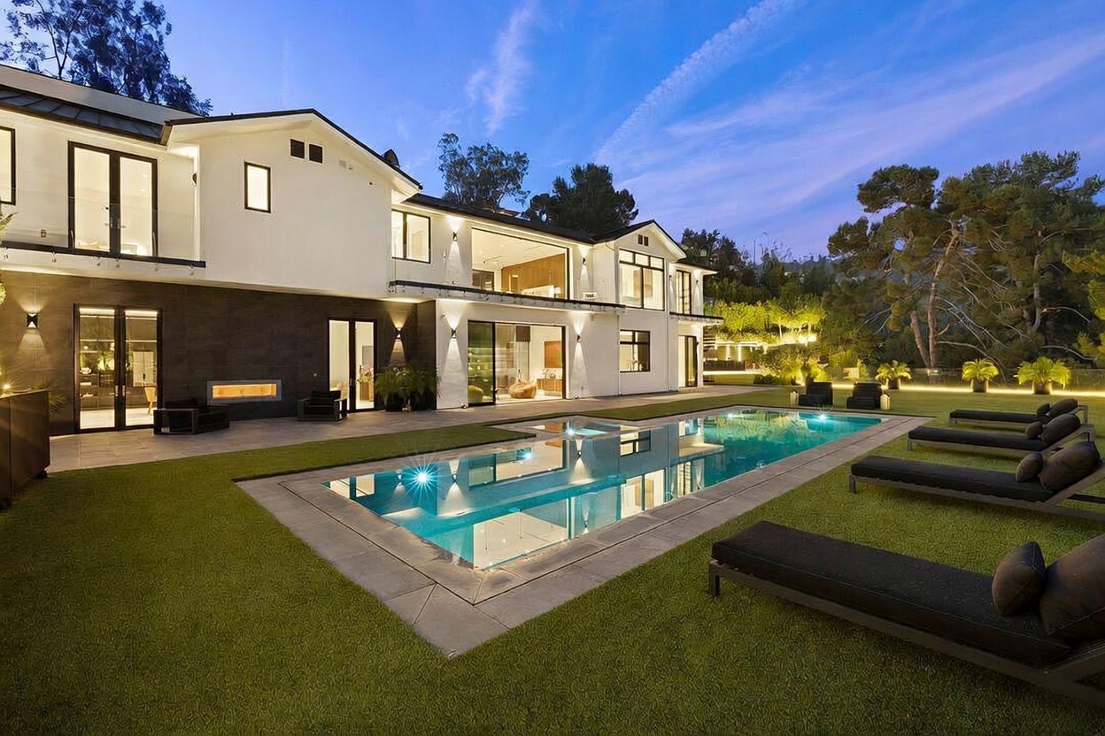 This modern luxury residence features a striking rear facade with a combination of white stucco and dark stone cladding. A rectangular swimming pool serves as the centerpiece of the backyard, surrounded by a manicured lawn and several lounge chairs. The home showcases large glass sliding doors that open to the patio, creating a seamless transition between indoor and outdoor living spaces.