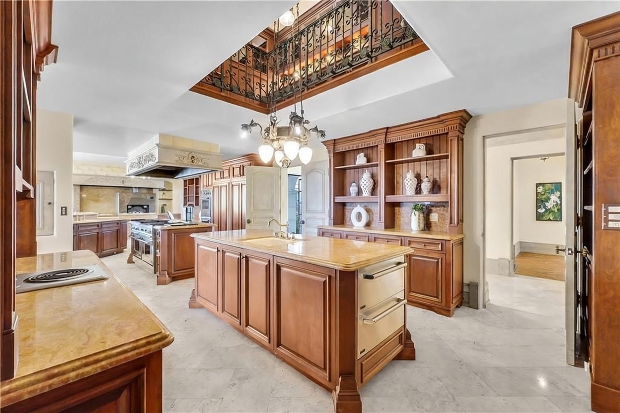 This expansive, traditional-style kitchen features a large central island with a stone countertop and integrated appliances. The space is defined by rich wood cabinetry, a decorative wrought-iron balcony overlooking the area from above, and a prominent chandelier. The perspective is a wide-angle shot from the kitchen entrance, showcasing the open layout and the transition into an adjacent hallway.