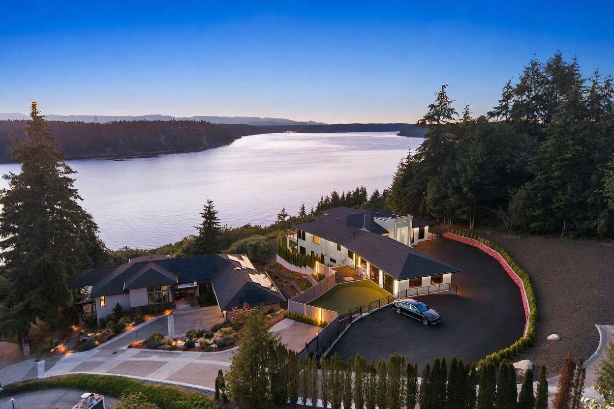 This high-angle aerial view captures a sprawling, modern luxury estate situated on a scenic waterfront bluff at twilight. The property features two distinct residential structures, expansive paved driveways, a manicured putting green, and lush surrounding forest, all overlooking a calm, wide river. The warm glow of the interior and exterior lighting contrasts beautifully with the cool blue tones of the evening sky and water.