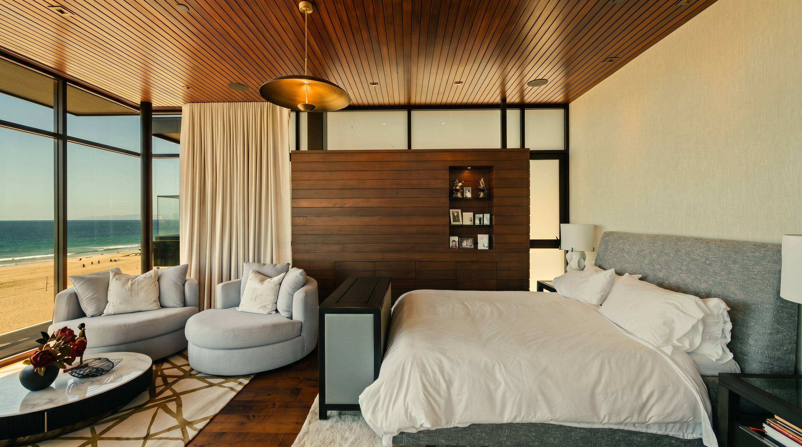 This luxurious primary bedroom features a striking wood-paneled ceiling and a matching wood partition wall that separates the sleeping area from a cozy sitting nook. Large windows provide an expansive, unobstructed view of the beach and ocean, filling the room with natural light. The space is furnished with a plush bed, a circular lounge chair, and a modern coffee table, creating a serene and sophisticated coastal retreat.