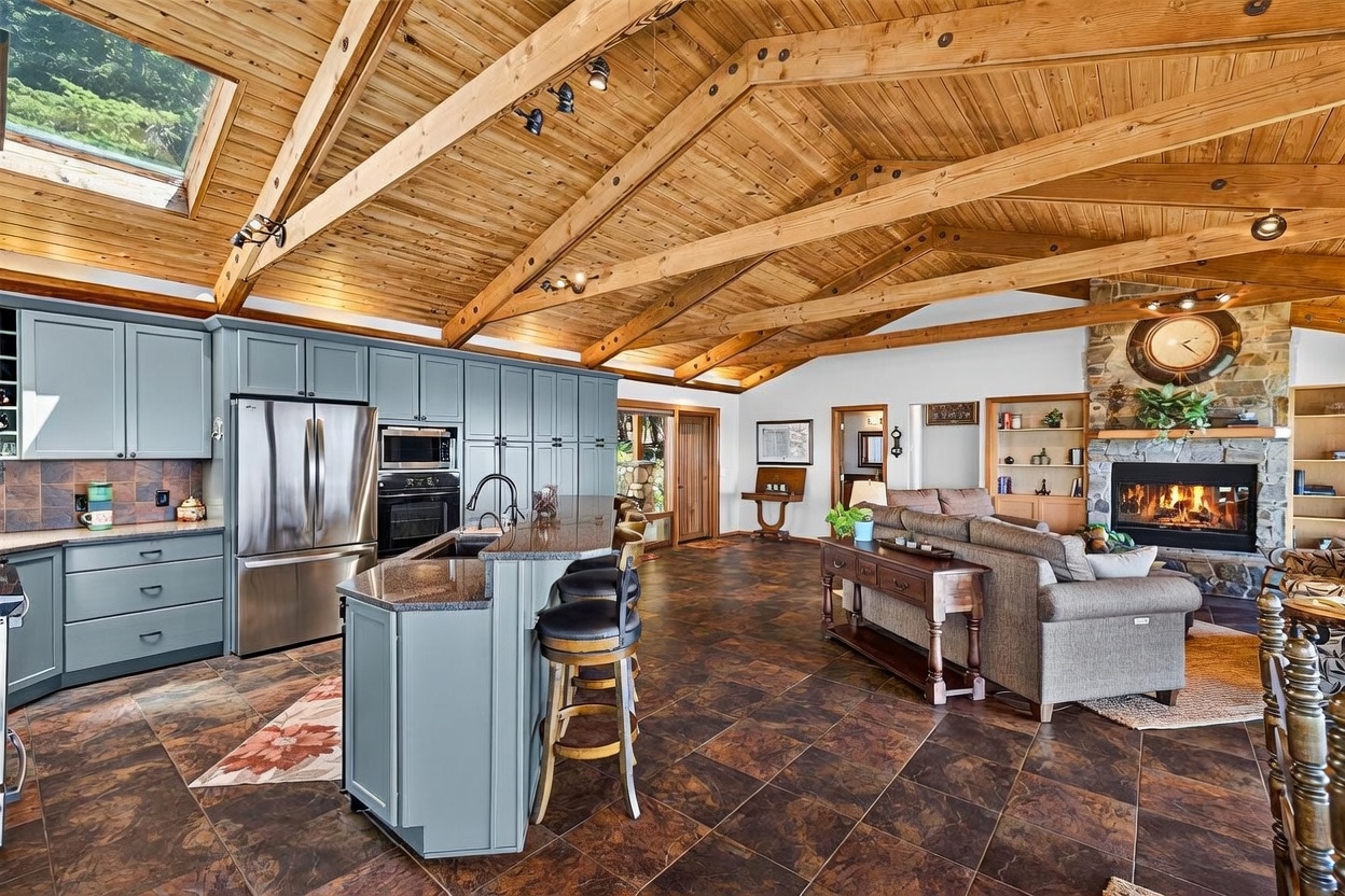 This open-concept living space features a spacious kitchen with blue cabinetry, a central island with bar seating, and dark patterned tile flooring. The vaulted ceiling is accented with exposed wooden beams and a large skylight, while the adjacent living area includes a stone-clad chimney breast with a glowing fireplace. The perspective captures the seamless flow between the culinary workspace and the cozy lounge, highlighting the rustic, warm atmosphere of the home.