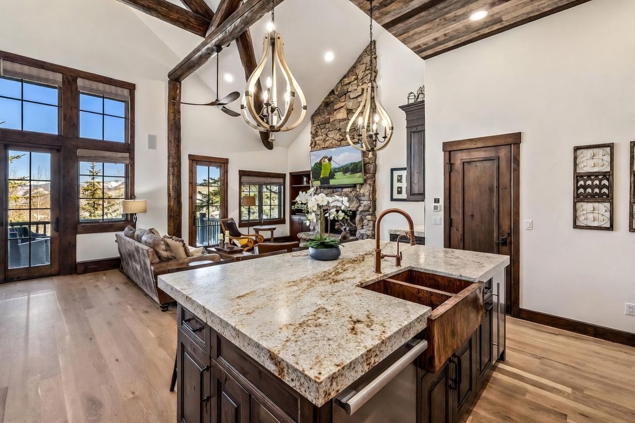 This open-concept kitchen features a large central island with a stone countertop and a copper farmhouse sink, serving as the focal point of the space. The room flows seamlessly into a living area characterized by vaulted ceilings with exposed timber beams, a stone-clad chimney breast, and large windows that frame mountain views. The aesthetic is rustic and refined, utilizing dark wood cabinetry and warm lighting to create an inviting, high-end atmosphere.