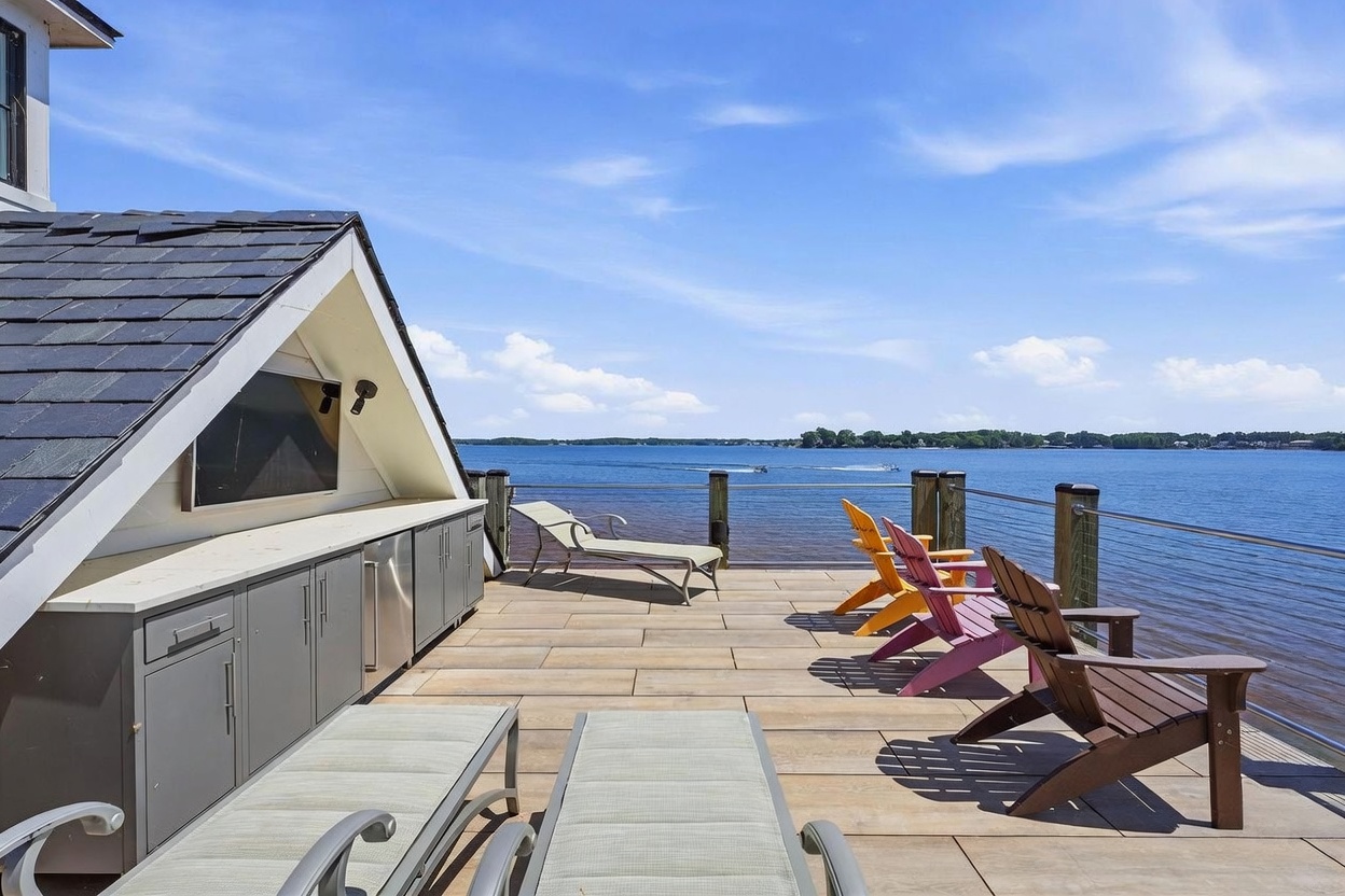 This expansive outdoor deck overlooks a large body of water, featuring a built-in outdoor kitchen area with gray cabinetry and a mounted television. The deck is furnished with several lounge chairs and colorful Adirondack chairs, providing a perfect space for relaxation and entertaining. The scene is captured from a slightly elevated perspective, emphasizing the open-air design and the scenic waterfront setting.