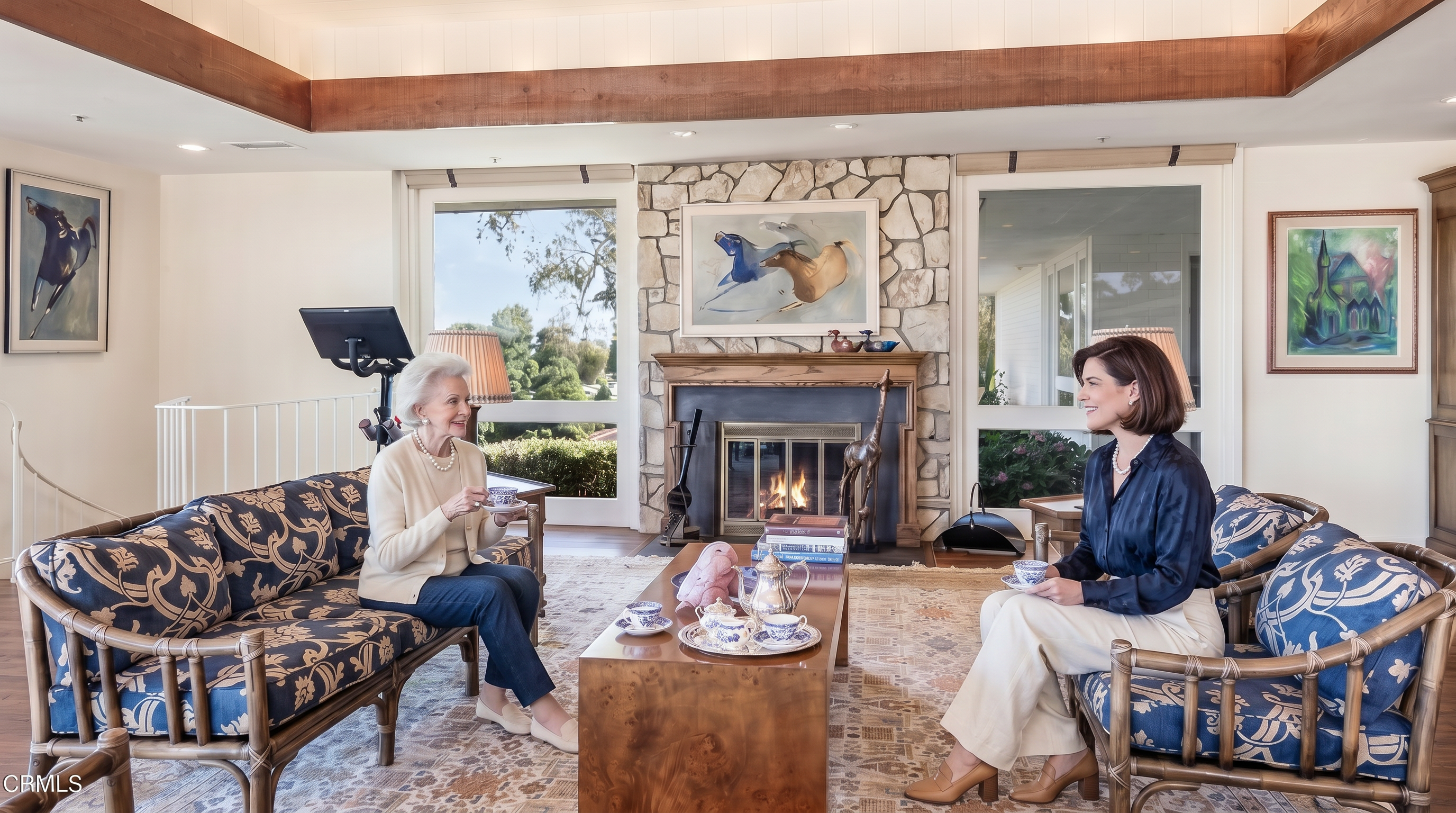 This spacious living room features a traditional aesthetic with a prominent stone-clad chimney breast and a wooden mantelpiece as the central focal point. The room is furnished with patterned blue and gold seating, a large wooden coffee table, and a detailed area rug, all set against a backdrop of hardwood flooring and large windows that invite natural light. A wooden beam ceiling detail adds architectural character to the space, which also includes a stationary exercise bike positioned near the window.