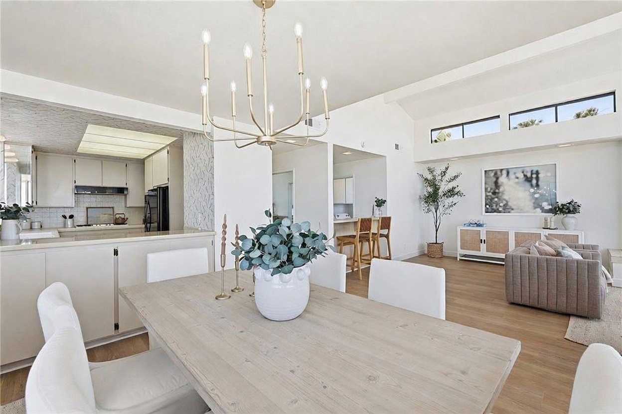 This bright, open-concept dining area features a light wood table as the centerpiece, illuminated by a modern multi-arm chandelier. The space flows seamlessly into a kitchen with white cabinetry and a living room area accented by a large piece of wall art and a neutral-toned sofa. Large windows near the ceiling line allow natural light to fill the room, creating an airy and inviting atmosphere.