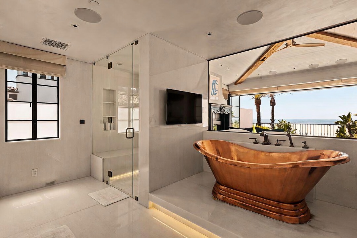 This luxurious primary bathroom features a striking copper soaking tub positioned on a raised platform, offering a direct view of the ocean through a large window. To the left, a glass-enclosed shower with built-in shelving provides a clean, modern aesthetic, while a wall-mounted television is positioned above the tub area. The space is finished with light-toned surfaces and recessed lighting, creating a serene and high-end atmosphere.