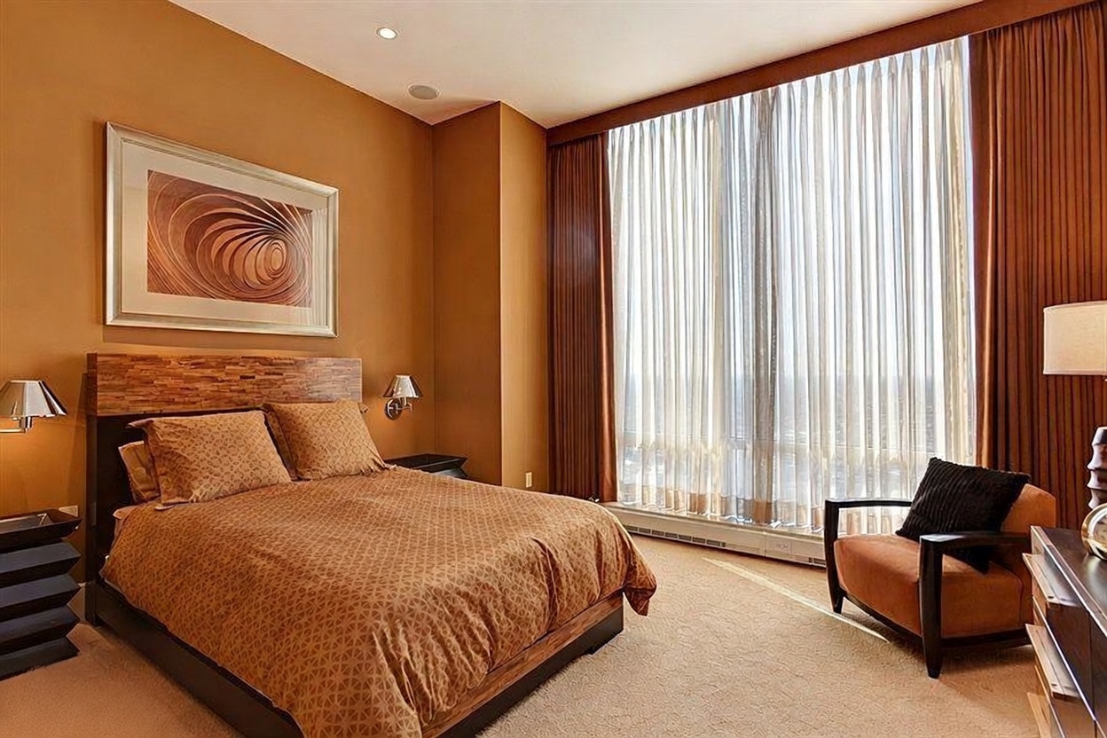 This primary bedroom features a warm, monochromatic color palette with rich orange-toned walls and matching bedding. A large bed with a textured wood headboard serves as the focal point, complemented by a modern armchair and floor-to-ceiling windows draped in sheer curtains. The room is illuminated by soft, warm lighting from wall-mounted lamps, creating a cozy and sophisticated atmosphere.