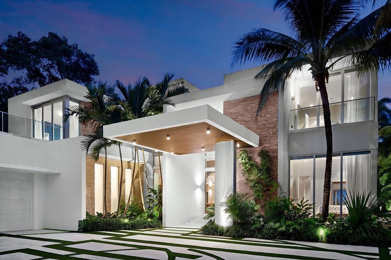 This striking modern residence features a dramatic, cantilevered entryway canopy with recessed lighting and a warm wood-paneled ceiling. The facade combines clean white architectural lines with textured stone cladding and expansive glass panels that offer a glimpse into the interior. Lush tropical landscaping, including palm trees and manicured greenery, frames the paved driveway, creating a sophisticated and inviting curb appeal at dusk.