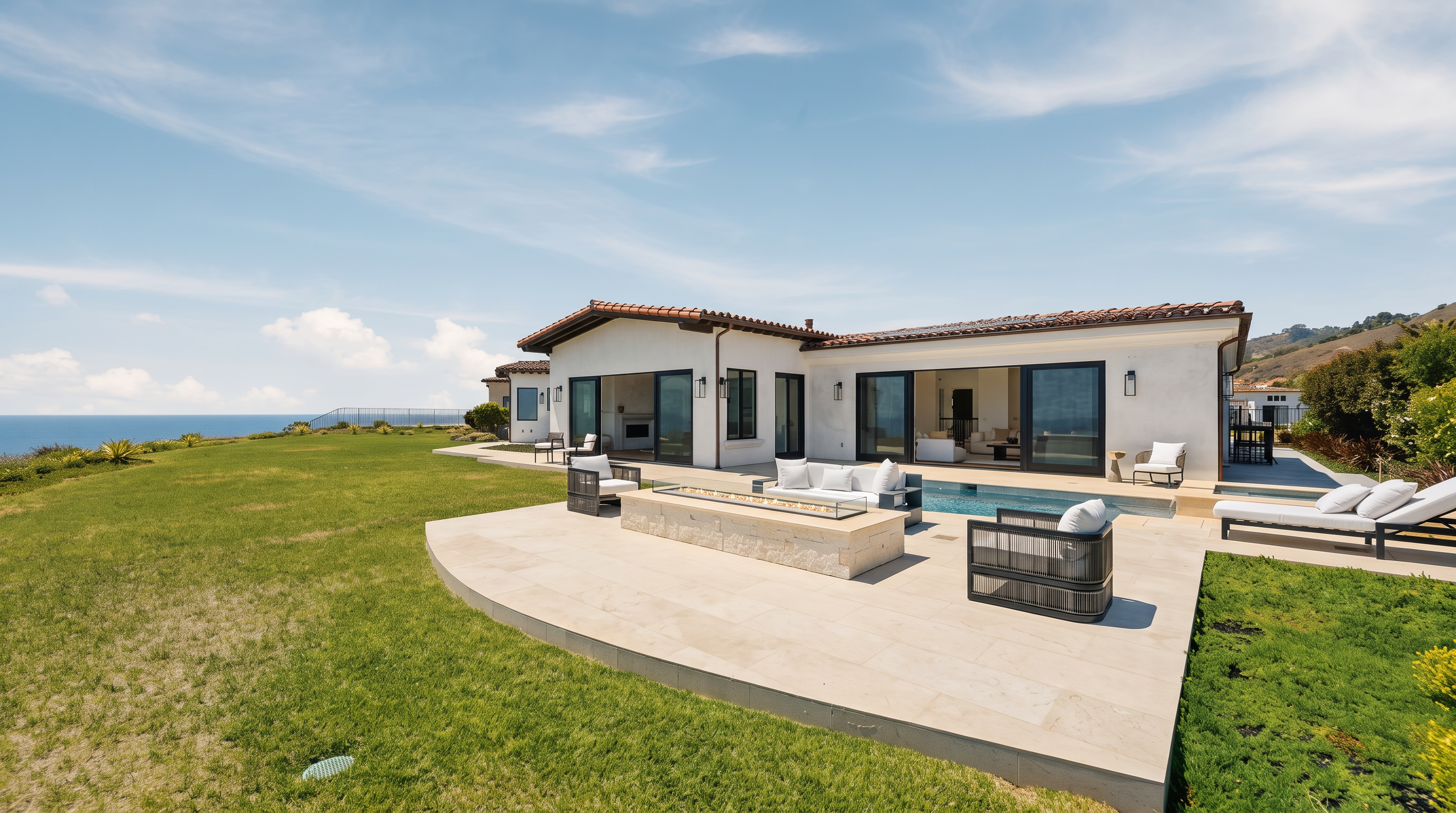 This expansive rear view of a luxury coastal residence showcases a seamless transition between indoor and outdoor living spaces. The property features a stone-paved patio with a central fire pit, a swimming pool, and large sliding glass doors that open to the interior. The home is set against a backdrop of a vast, manicured lawn and a panoramic ocean view under a clear blue sky.