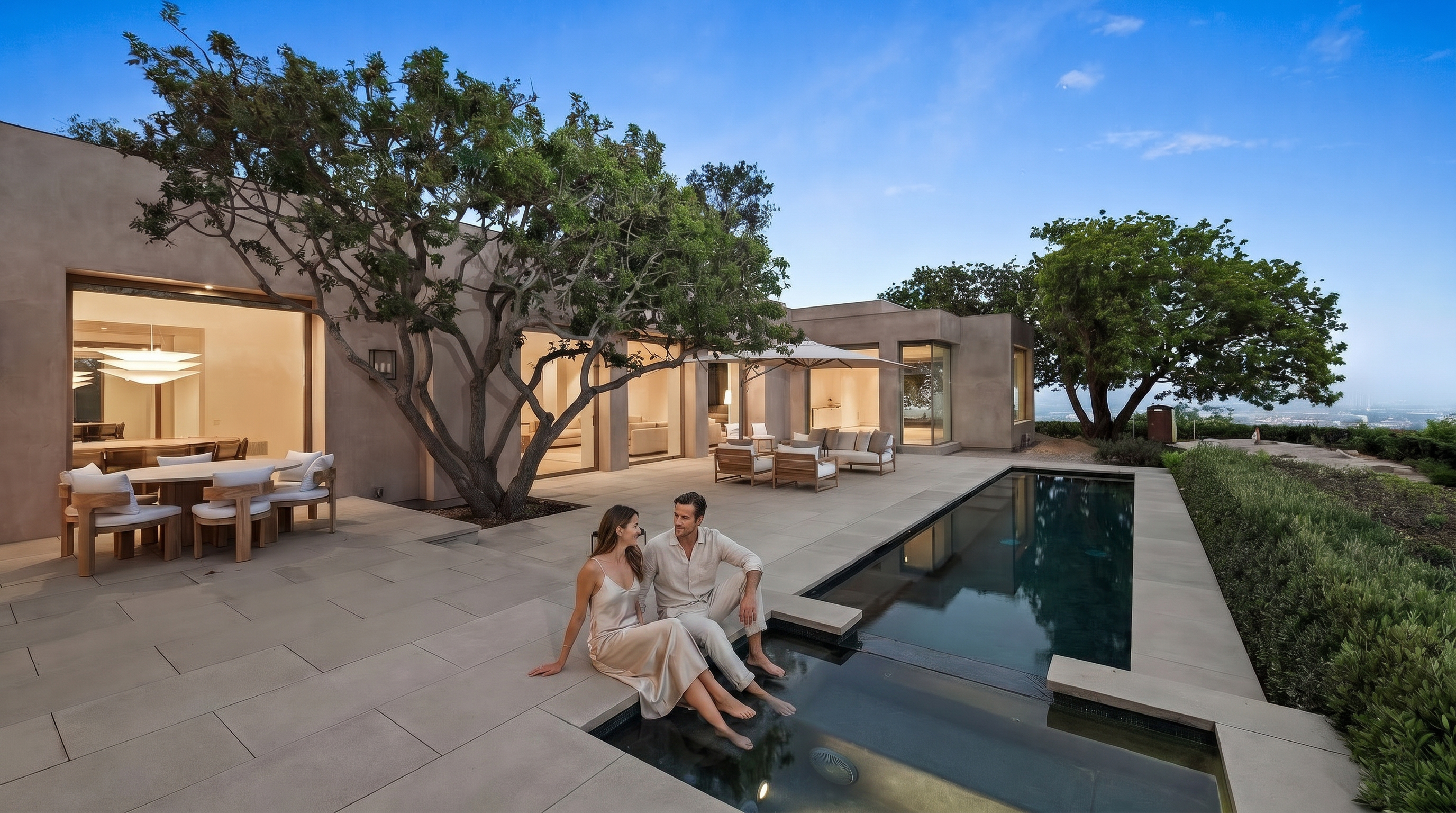 This stunning contemporary outdoor living space features a sleek, rectangular lap pool integrated into a spacious stone patio. The area is furnished with modern outdoor dining and lounge sets, shaded by a large tree and a patio umbrella, creating a perfect environment for relaxation and entertaining. The architectural design emphasizes clean lines and seamless indoor-outdoor flow, with expansive glass doors opening into the home's interior.