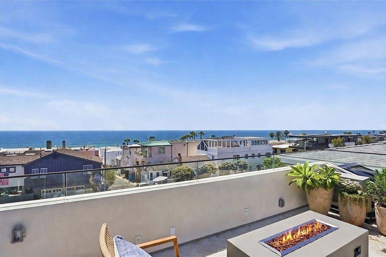 This elevated balcony offers a panoramic, unobstructed view of the ocean and the surrounding coastal neighborhood. The space is furnished with a modern, rectangular fire pit and a comfortable chair, creating an inviting outdoor lounge area. A sleek glass railing ensures the view remains the focal point, while potted plants add a touch of greenery to the clean, contemporary aesthetic.