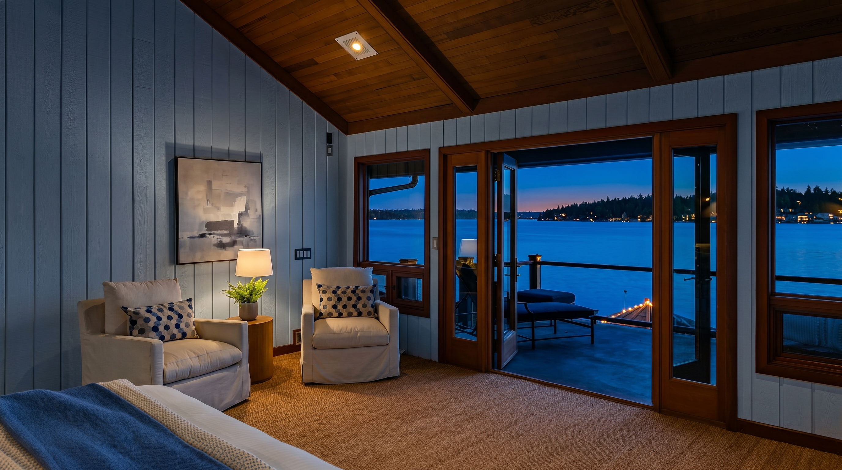 This serene primary bedroom features a vaulted wood-paneled ceiling and white vertical shiplap walls, creating a warm, coastal-inspired atmosphere. The room is anchored by two comfortable armchairs and opens directly onto a private deck through large glass sliding doors, offering a stunning, unobstructed view of the lake at sunset. The neutral color palette and natural textures, such as the woven rug, emphasize a tranquil and luxurious living space.