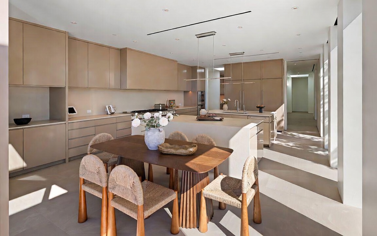 This sophisticated, modern kitchen features a seamless blend of warm, neutral tones and minimalist design, highlighted by sleek cabinetry and a large central island. A wooden dining table with woven chairs sits in the foreground, creating a cozy yet elegant eating area that flows directly into the culinary space. The open-concept layout is bathed in natural light, emphasizing the high-end finishes and clean architectural lines.
