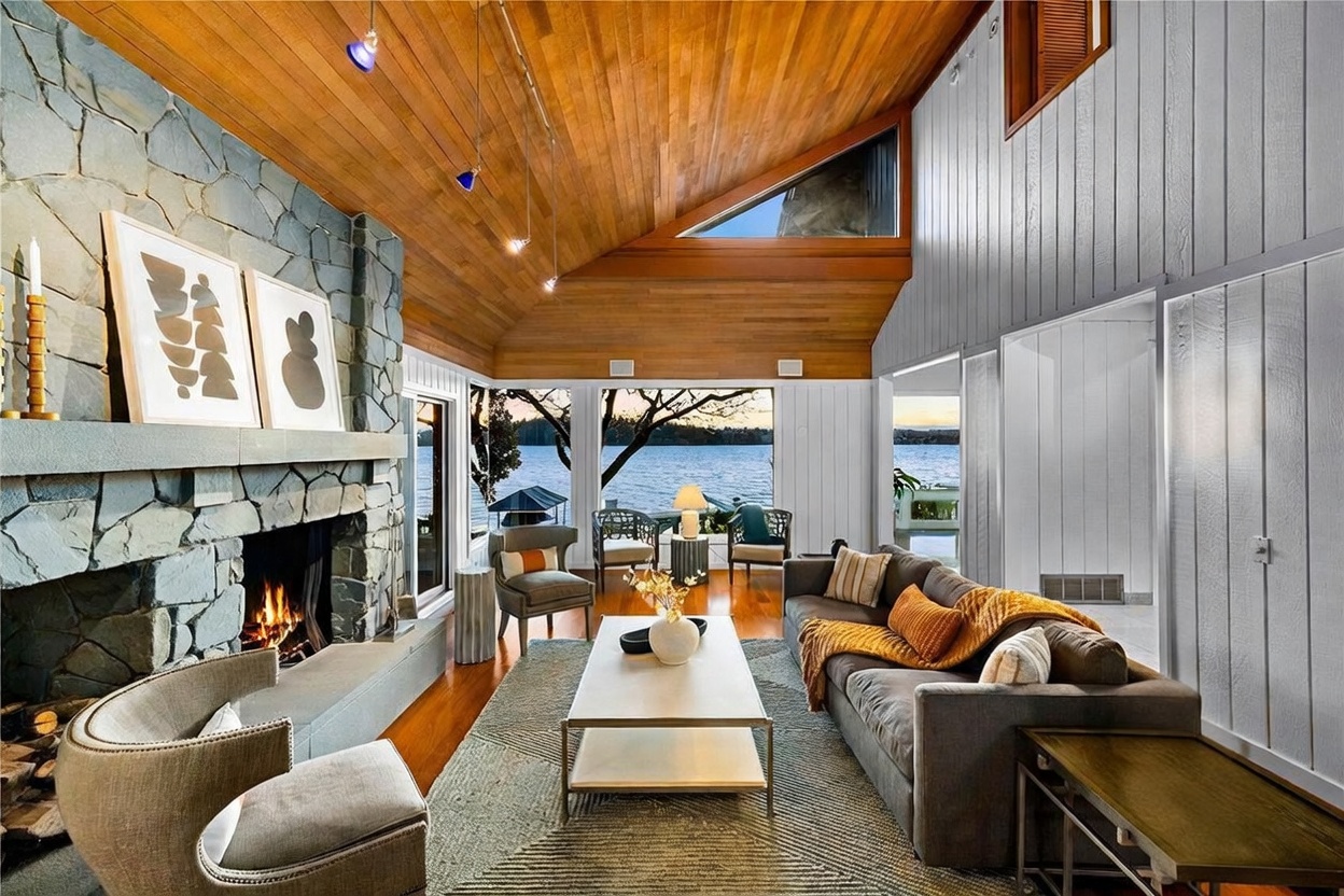 This stunning living room features a dramatic vaulted wood-paneled ceiling and a massive floor-to-ceiling stone fireplace that serves as the room's focal point. Large windows provide an expansive, unobstructed view of the lake, bathing the space in natural light and creating a seamless connection to the outdoors. The room is furnished with a comfortable grey sofa and stylish accent chairs, blending rustic textures with modern, sophisticated design.