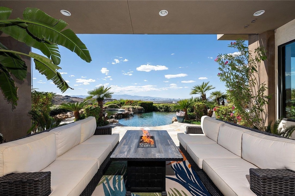 This covered patio offers a luxurious outdoor living space featuring two large, dark-wicker sofas with light-colored cushions arranged around a central fire table. The space opens directly to a landscaped backyard with a custom swimming pool and a panoramic view of the distant city skyline. Lush tropical plants and palm trees frame the scene, creating a serene and upscale atmosphere perfect for entertaining.