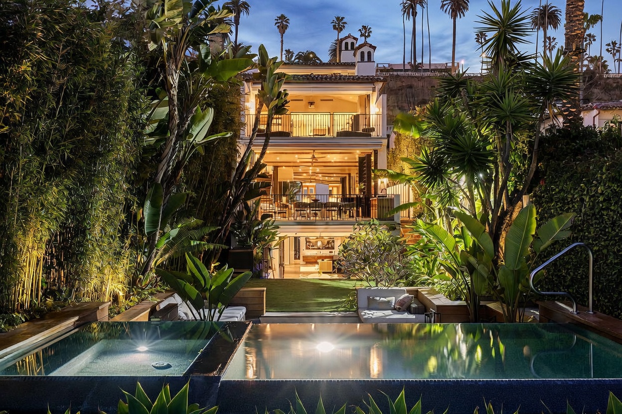 This rear view of a multi-story luxury residence showcases a tiered architectural design with expansive balconies and large glass doors that open to the outdoors. The property features a private swimming pool and spa in the foreground, surrounded by lush tropical landscaping and mature palm trees. The warm evening lighting creates an inviting and sophisticated atmosphere, highlighting the seamless transition between the indoor living spaces and the private backyard oasis.