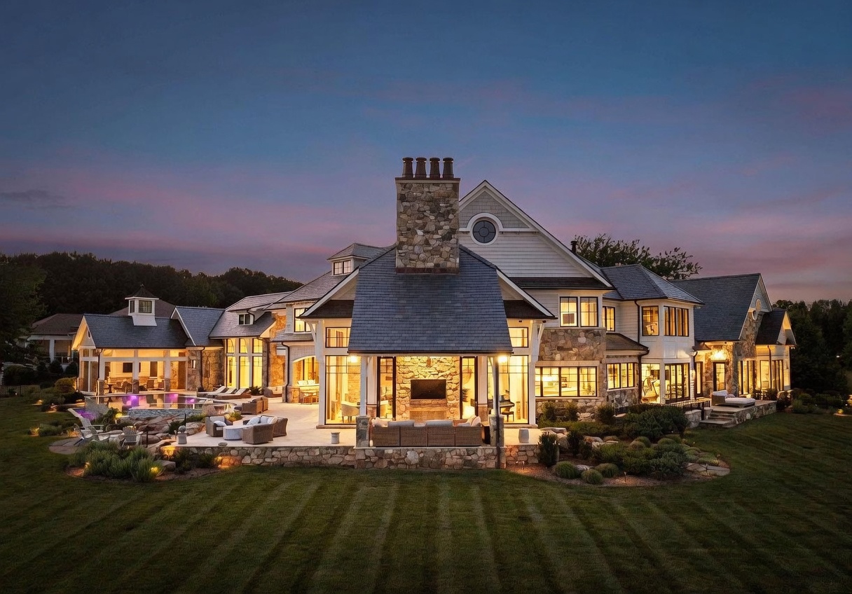 This expansive luxury estate is captured from a rear perspective at twilight, showcasing a multi-gabled roofline and a sophisticated blend of stone and siding. The home features a large stone-clad chimney breast, a covered outdoor living area with a mounted television, and an integrated swimming pool surrounded by a stone patio. The scene is illuminated by warm interior lighting and landscape accents, creating an inviting and high-end atmosphere against a soft sunset sky.