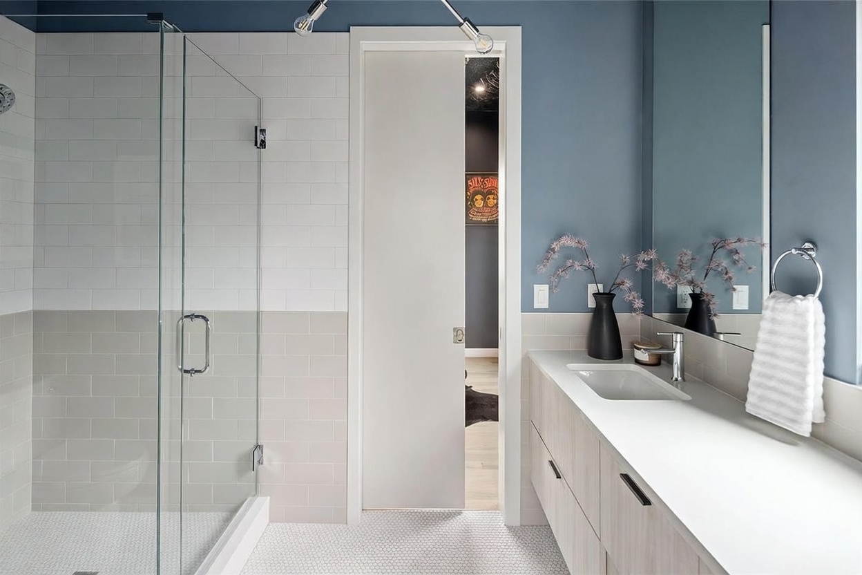 This modern bathroom features a sleek vanity with light wood cabinetry and a white countertop, complemented by a large mirror and a wall-mounted towel ring. To the left, a glass-enclosed shower is finished with a combination of white and neutral-toned rectangular tiles, while the floor is covered in small, circular mosaic tiles. A sliding door provides access to an adjacent room, creating a clean and contemporary aesthetic.
