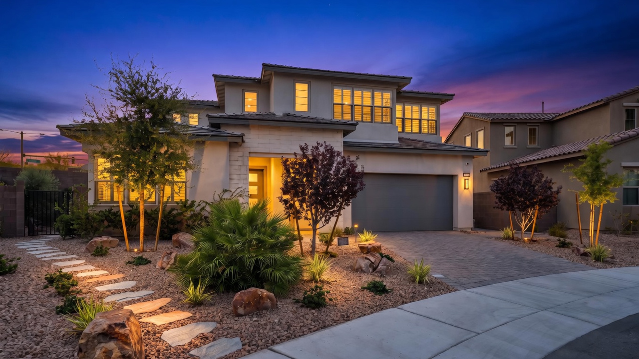 This modern two-story residence presents an inviting front facade at dusk, highlighted by warm landscape lighting and a stylish paver driveway. The exterior features a blend of stucco and stone textures, complemented by a desert-inspired xeriscape design with mature greenery. Its balanced architecture and well-lit entry create a sophisticated and welcoming curb appeal.