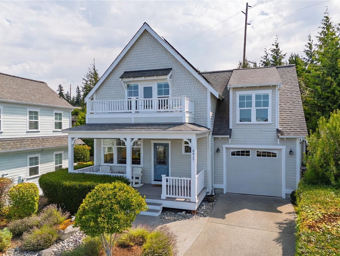 This charming two-story home features light-colored shingle and horizontal siding with a welcoming front porch and a second-story balcony. The property includes an attached garage, a concrete driveway, and a neatly landscaped front yard with mature shrubs and perennials. The architectural style is reminiscent of a coastal cottage, offering a bright and inviting curb appeal.